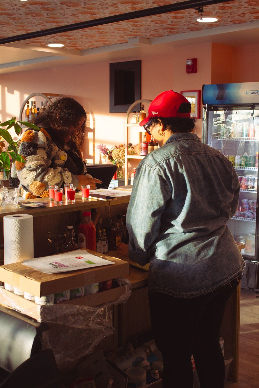 Two people at a cafe counter with bottles and refrigerator in warm sunlight.
