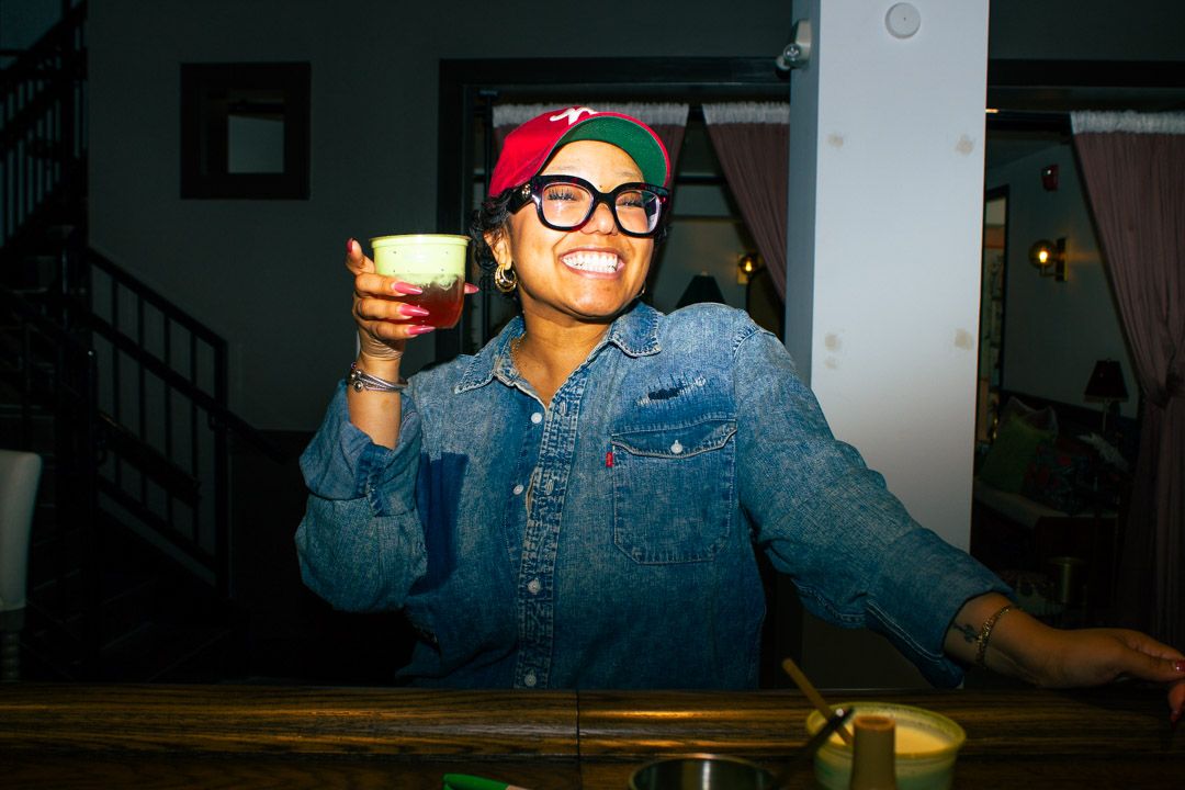 Smiling woman in denim shirt and colorful hat holding a red drink at a bar.