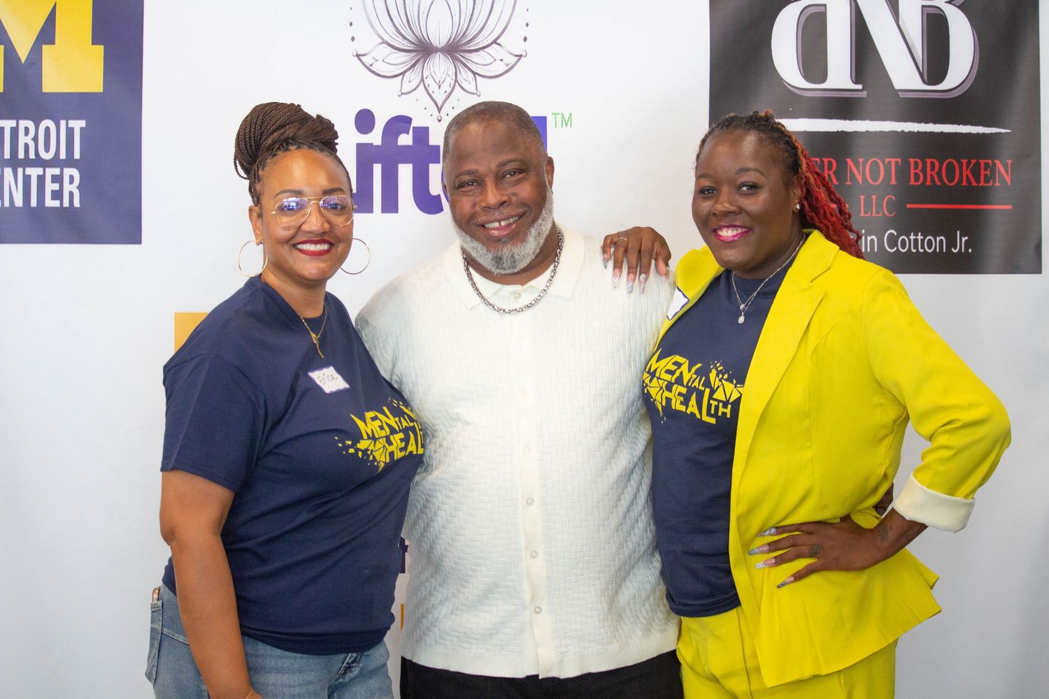 Three people smiling and posing together at a community health event in Detroit, with branded banners in the background.