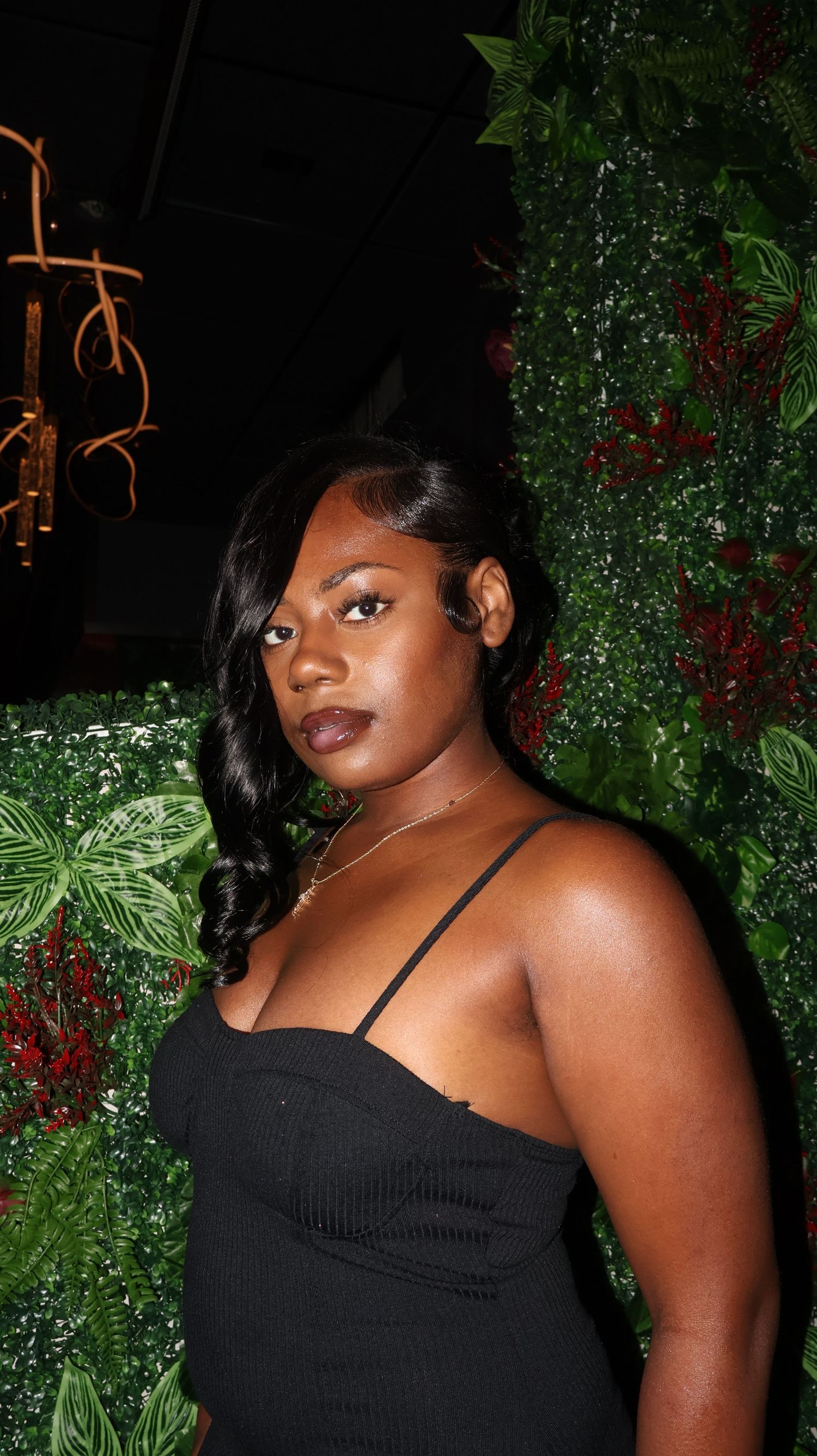 Young woman in black spaghetti strap dress poses before lush green floral wall at night event.