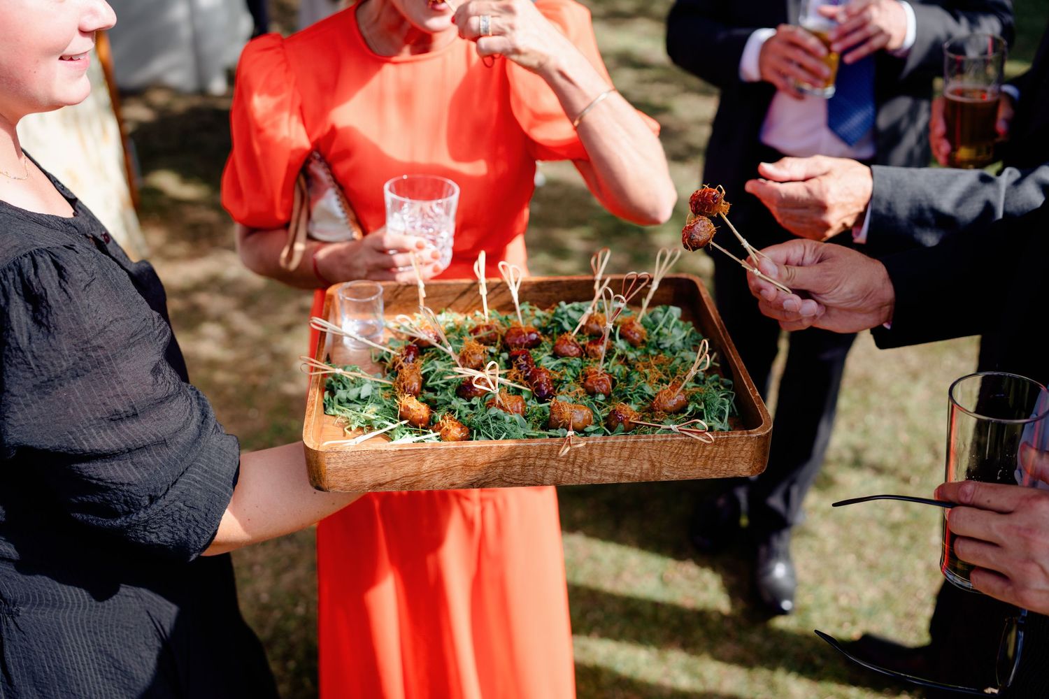 Wedding guest in red dress holds wooden tray of canapés with skewers at outdoor reception.