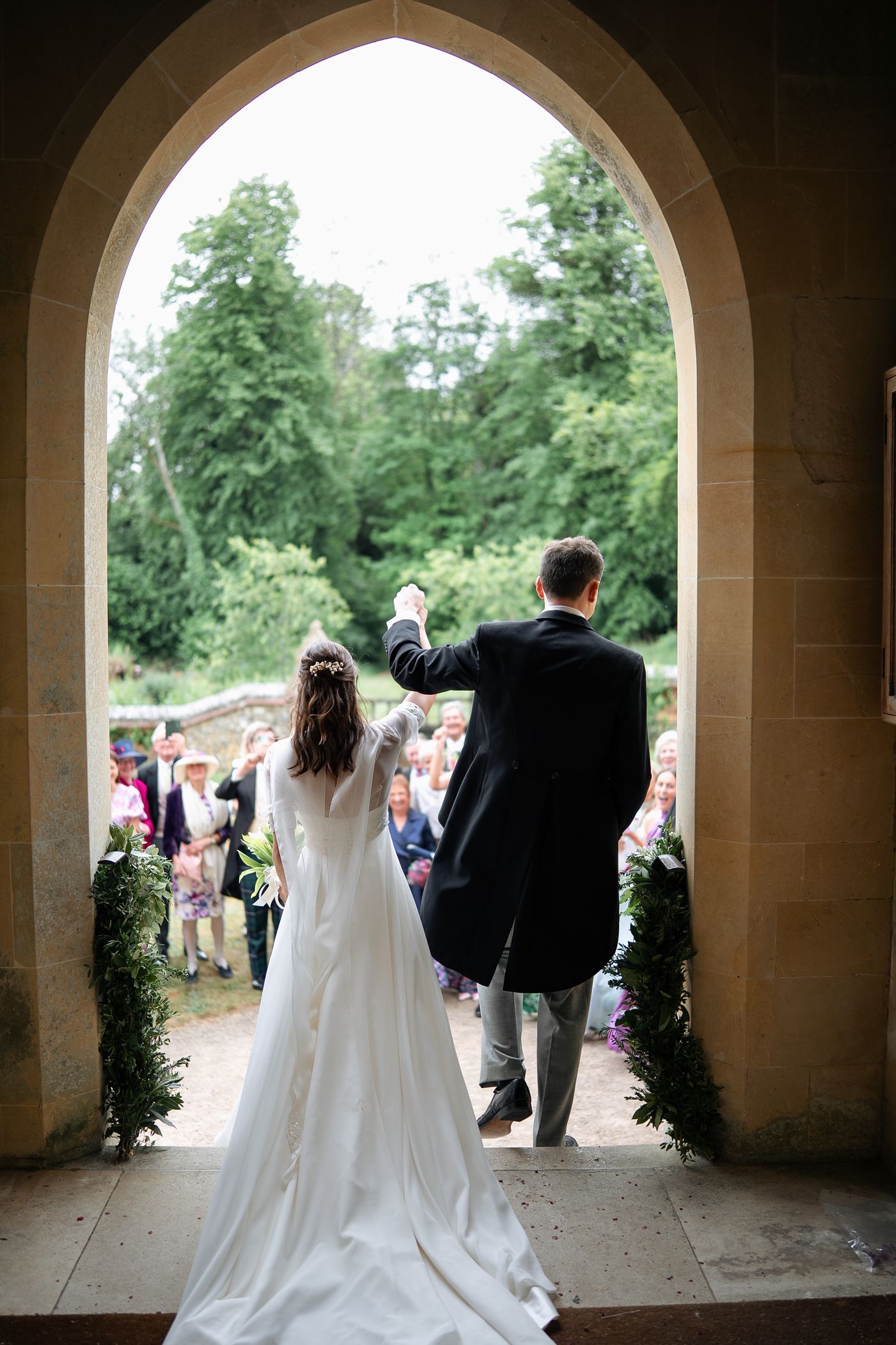 Bride and groom stand in a stone church archway raising hands as guests gather outside.