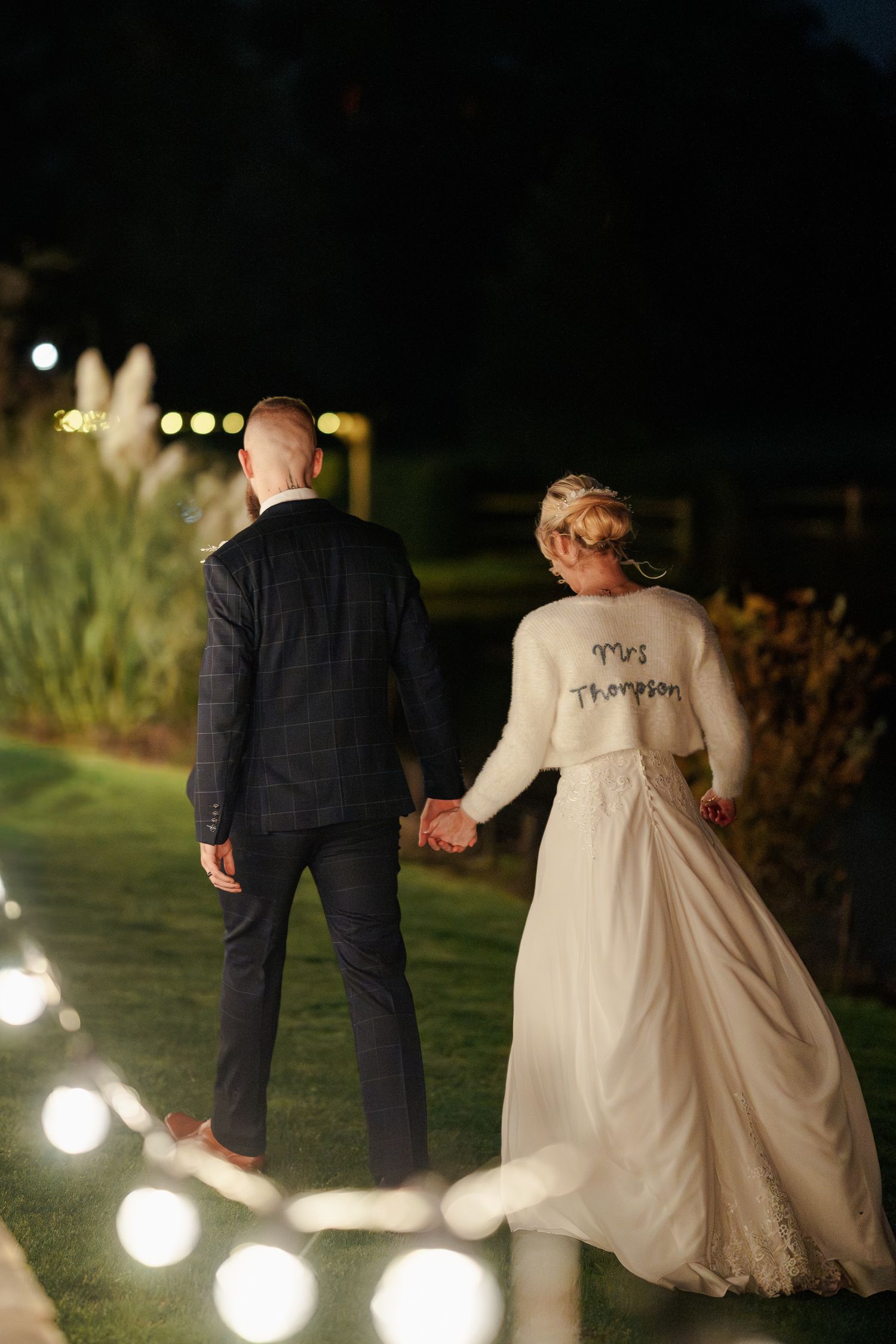 Bride wearing Mrs Thompson cardigan holds grooms hand walking along lit garden path.