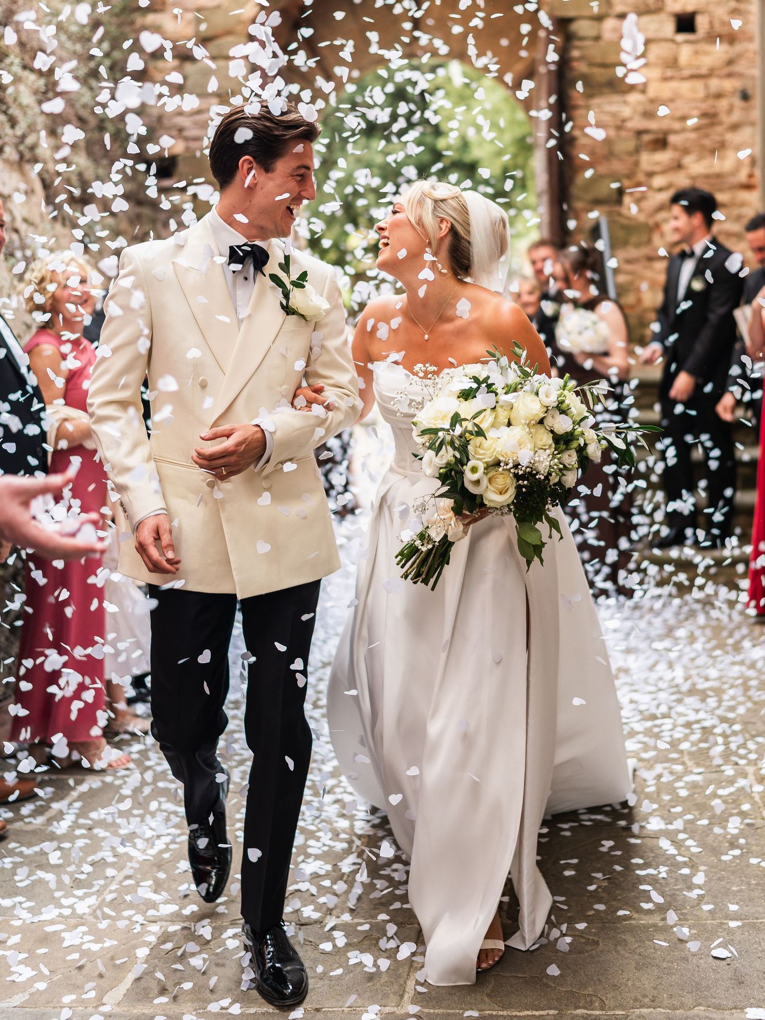 Elegant bride and groom walk through white confetti shower outside rustic stone castle wedding venue.