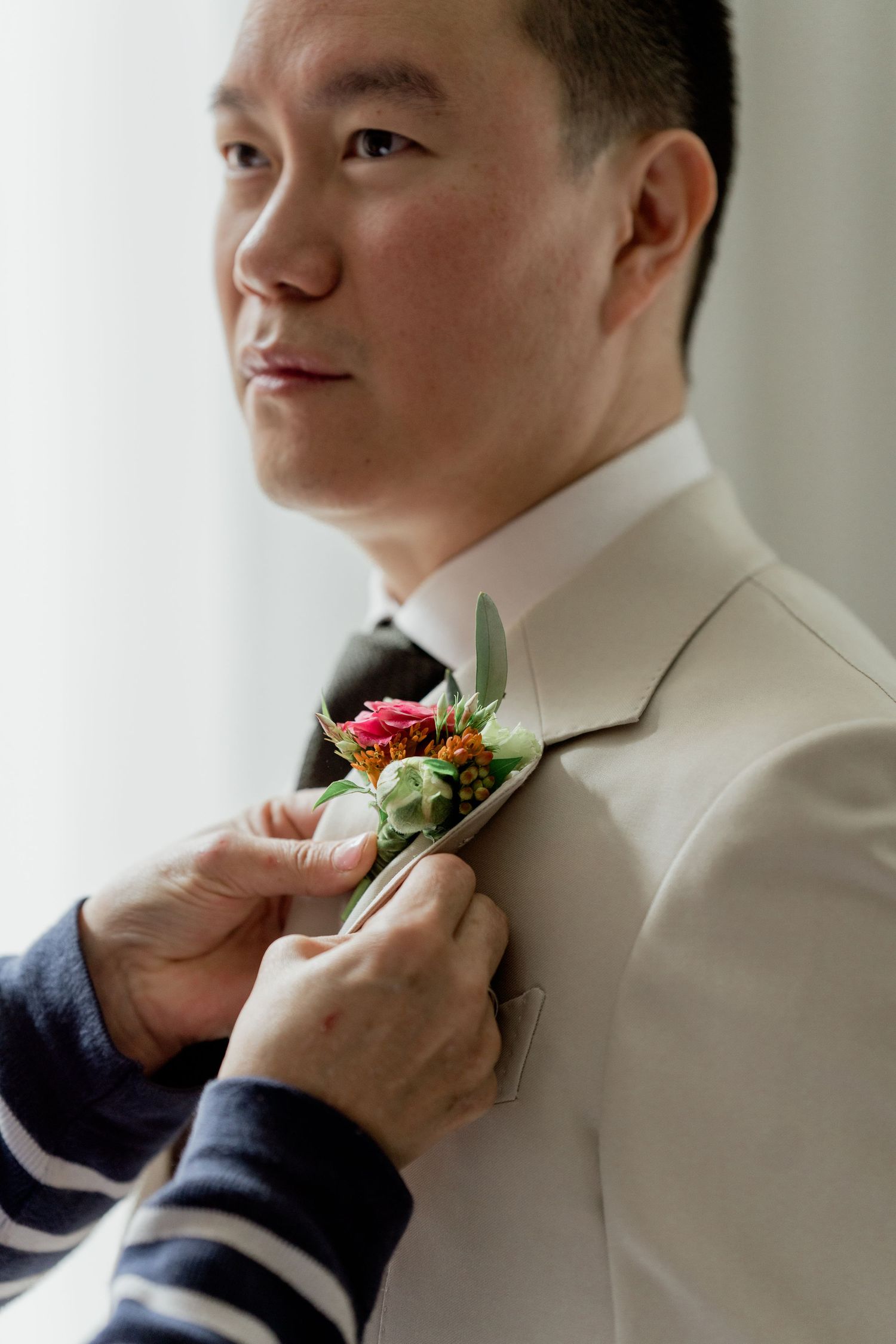 Groom in beige suit has colorful floral boutonniere pinned to lapel before wedding.