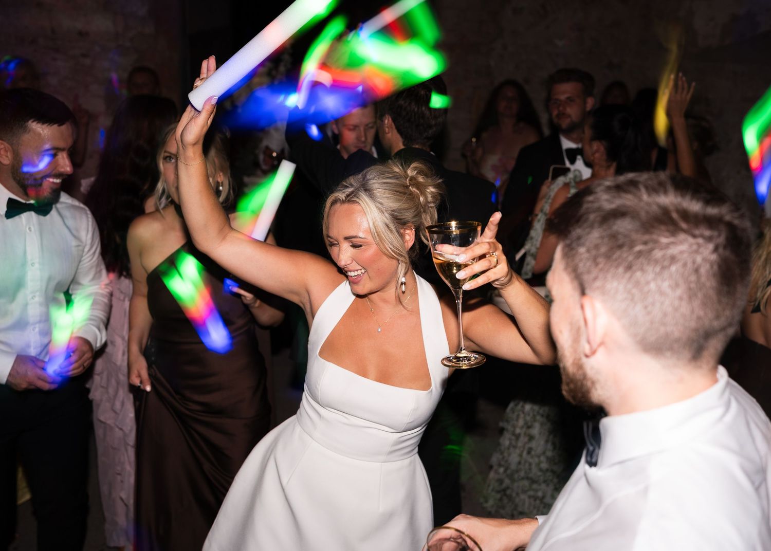 Bride dancing with champagne glass under colorful glow stick lights at a lively wedding reception.