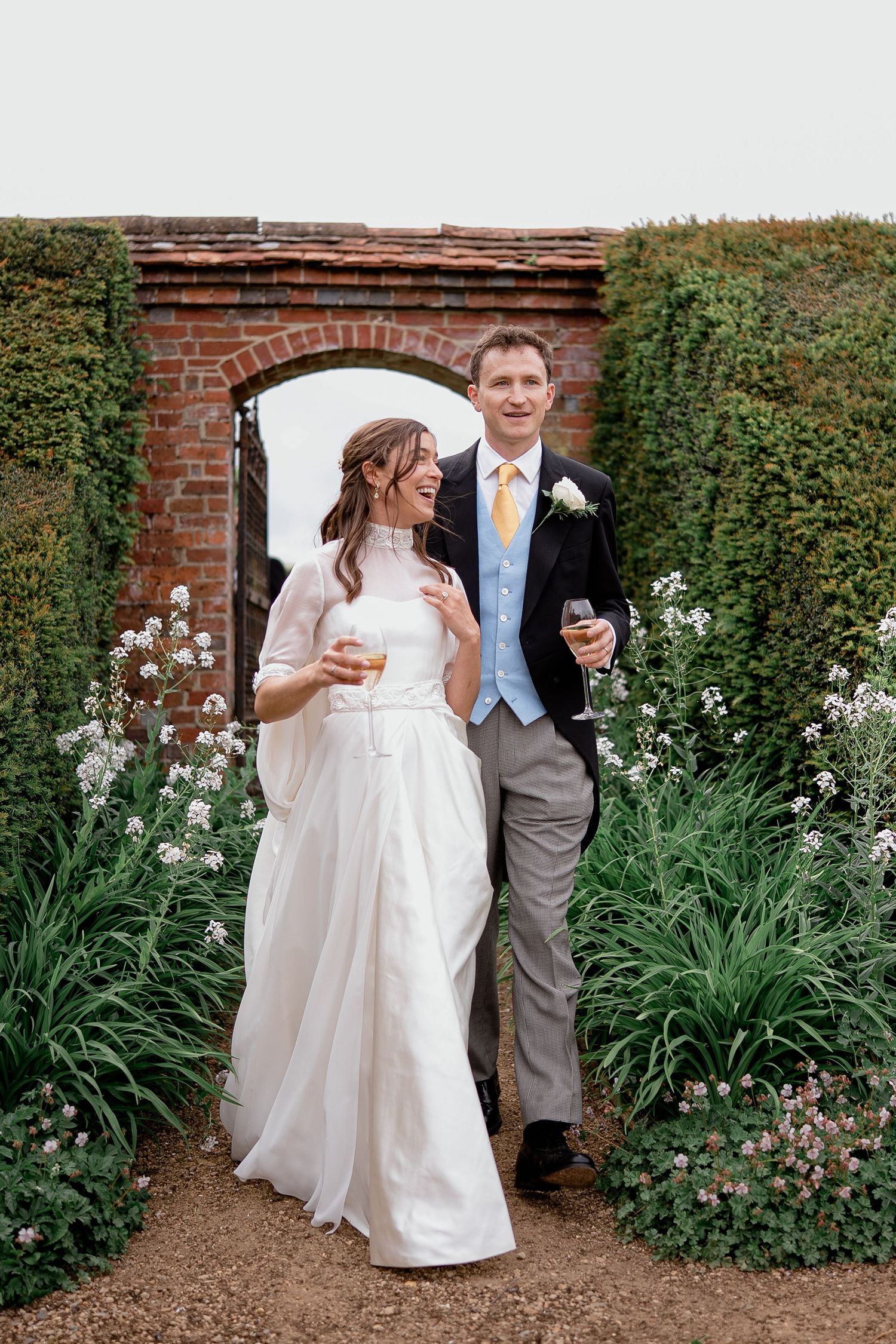 Bride and groom smiling together in a lush walled garden, holding drinks on their wedding day.