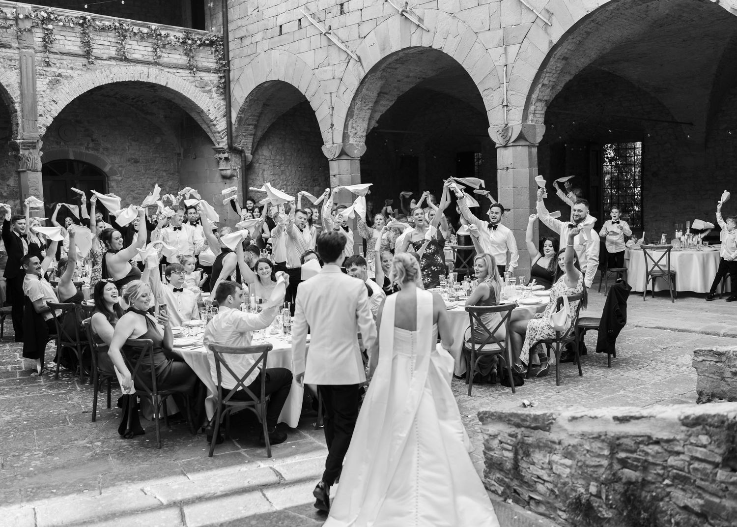 Black and white photo of wedding guests cheering as bride and groom enter elegant arched courtyard reception.