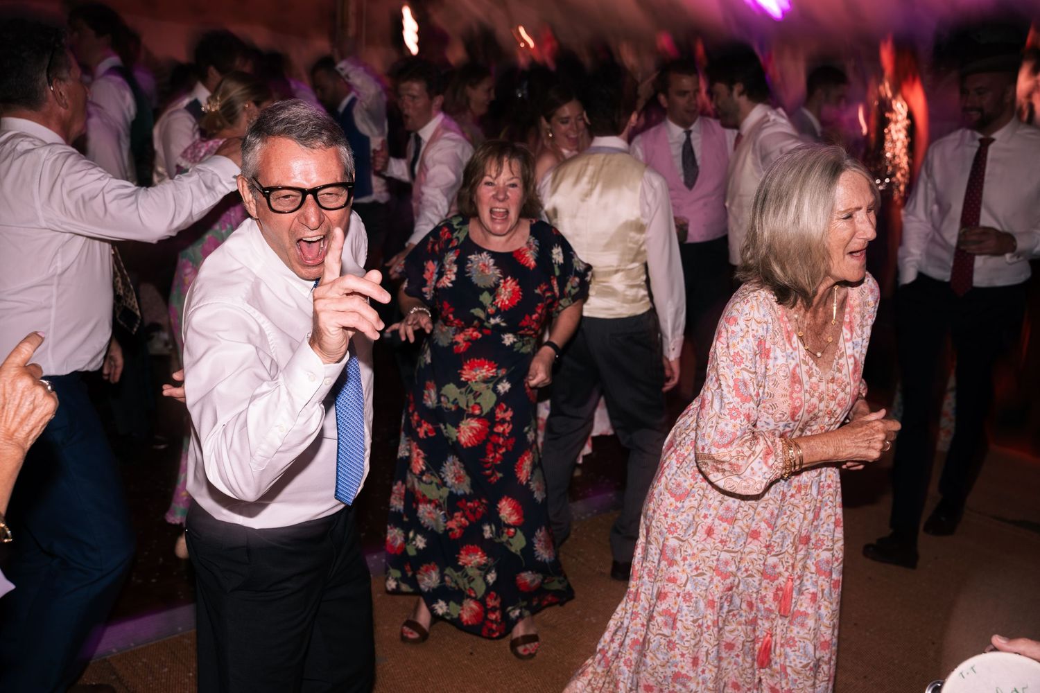 Older wedding guests dancing energetically on packed reception dance floor at night.