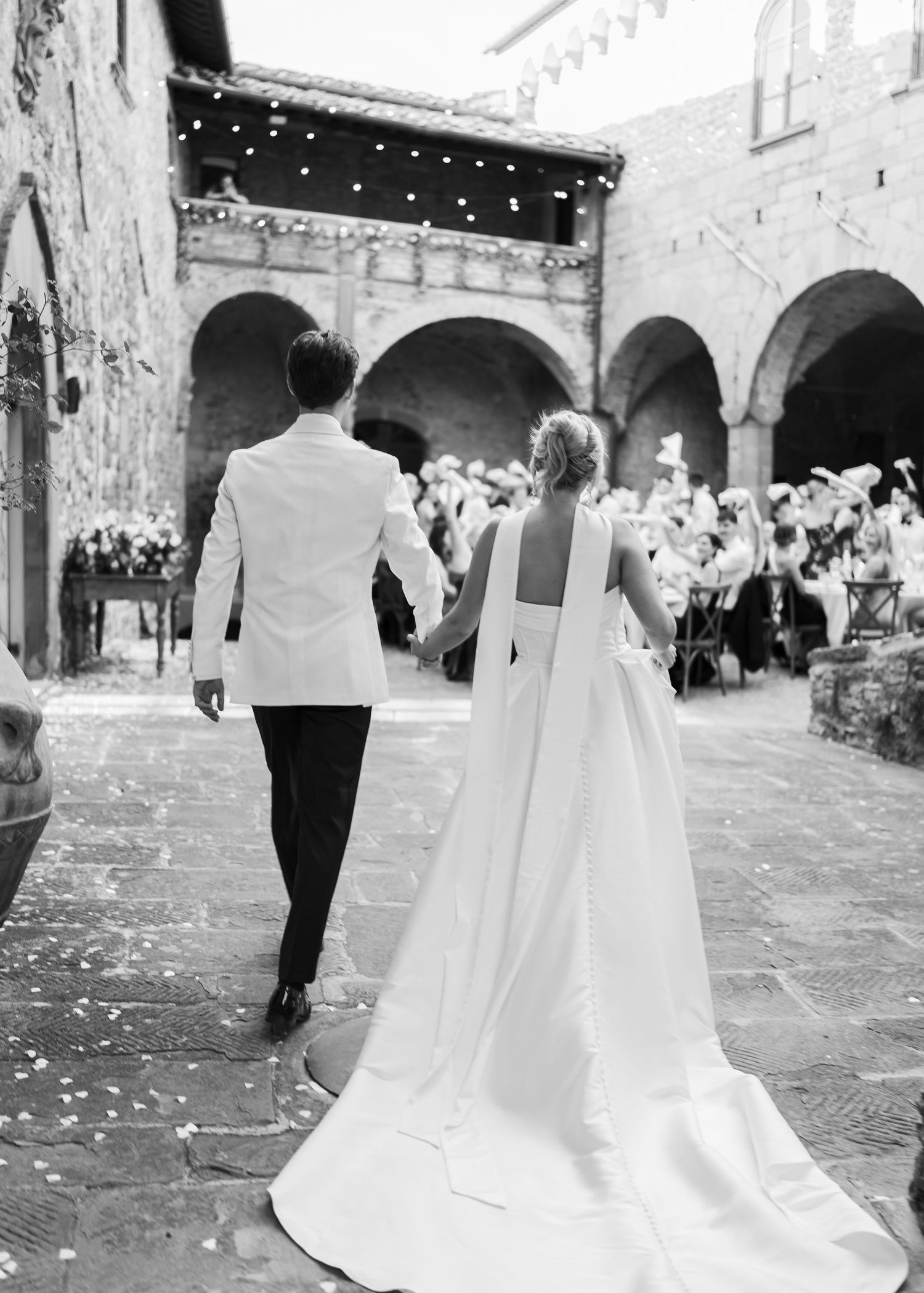 Black and white rear view of bride and groom walking into stone courtyard wedding reception venue.