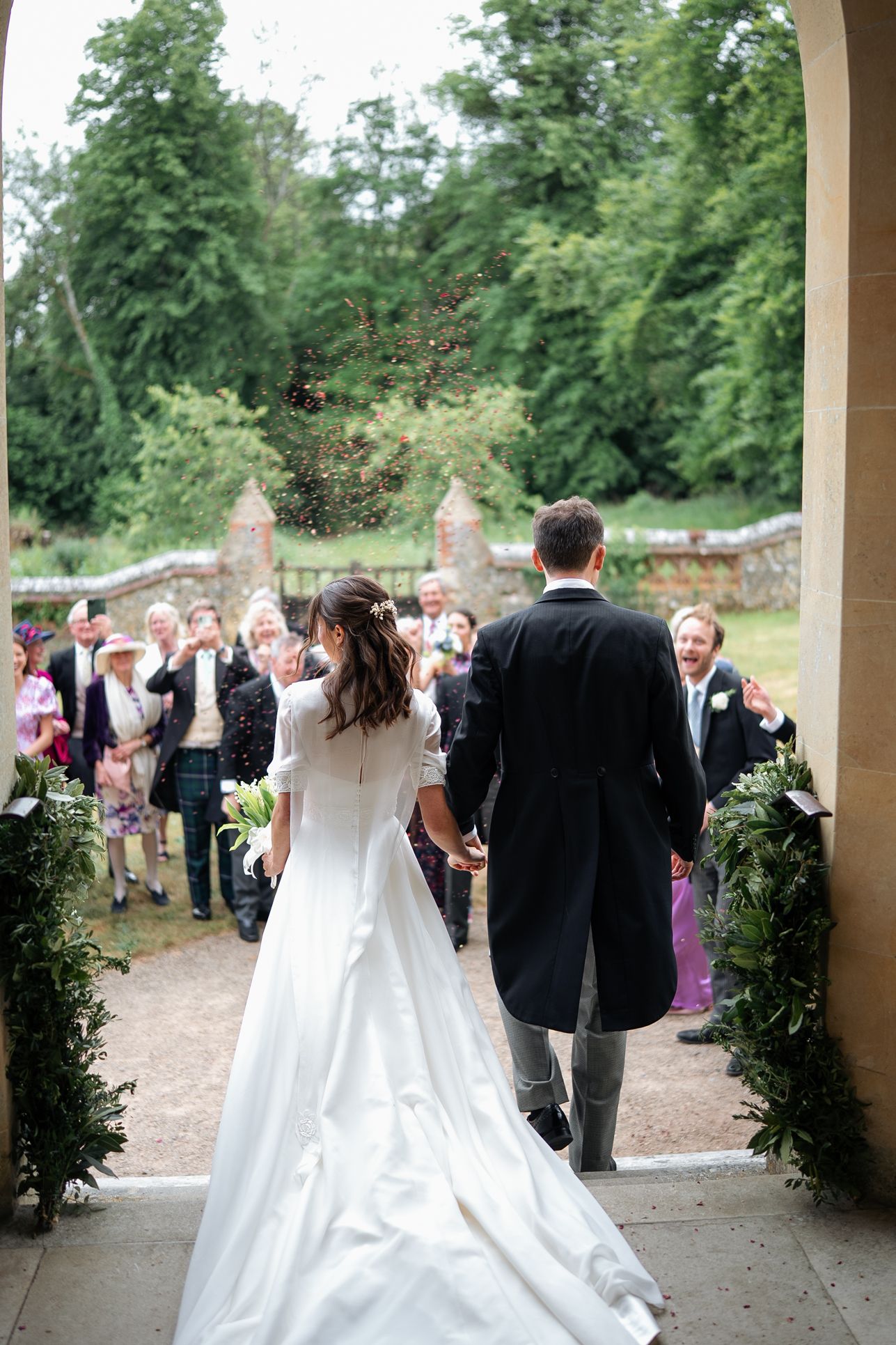 Newlyweds walk through archway as guests throw confetti at outdoor wedding celebration.