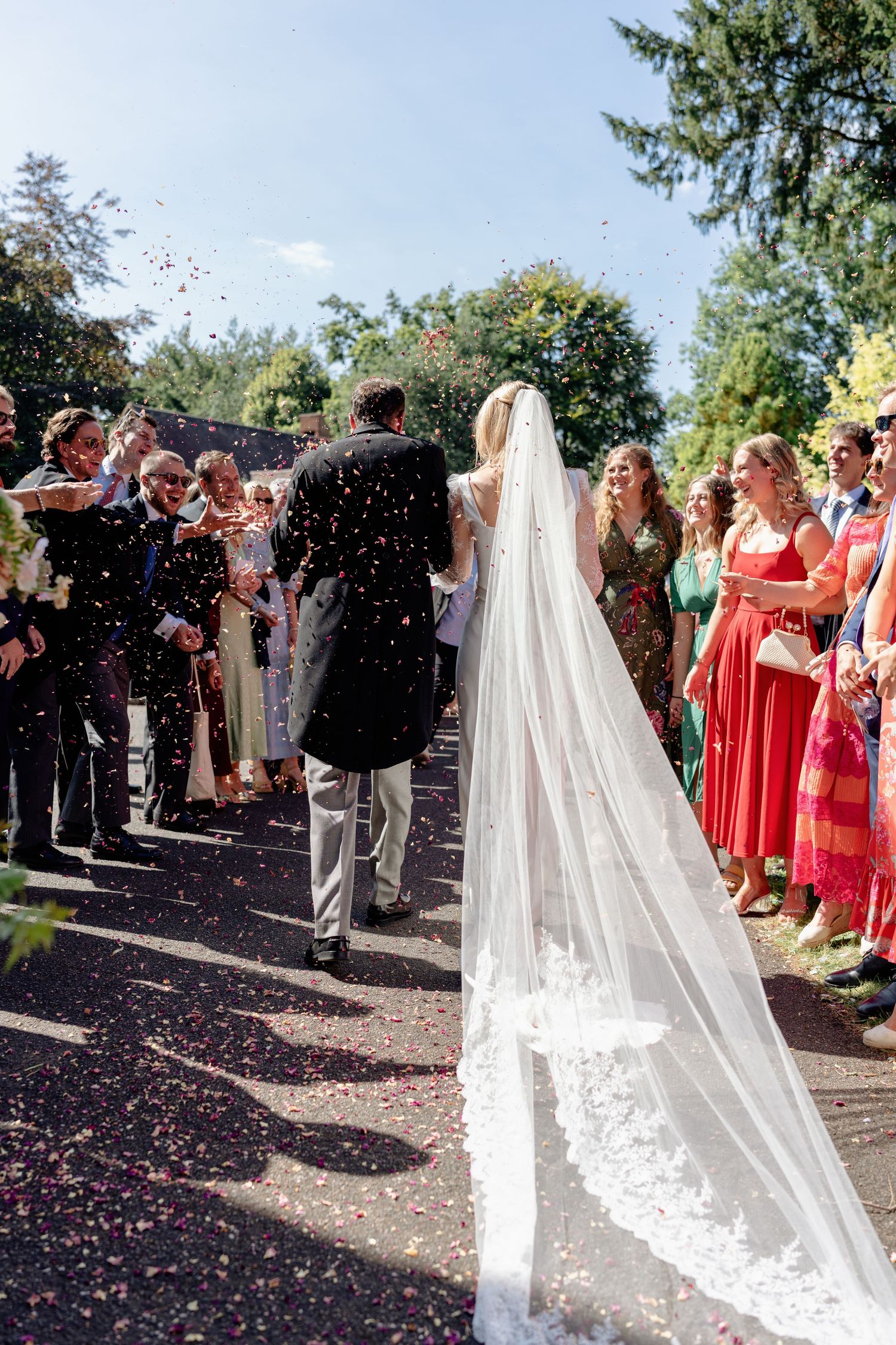 Bride in lace cathedral veil kissed by groom as colorful confetti falls during outdoor wedding ceremony.