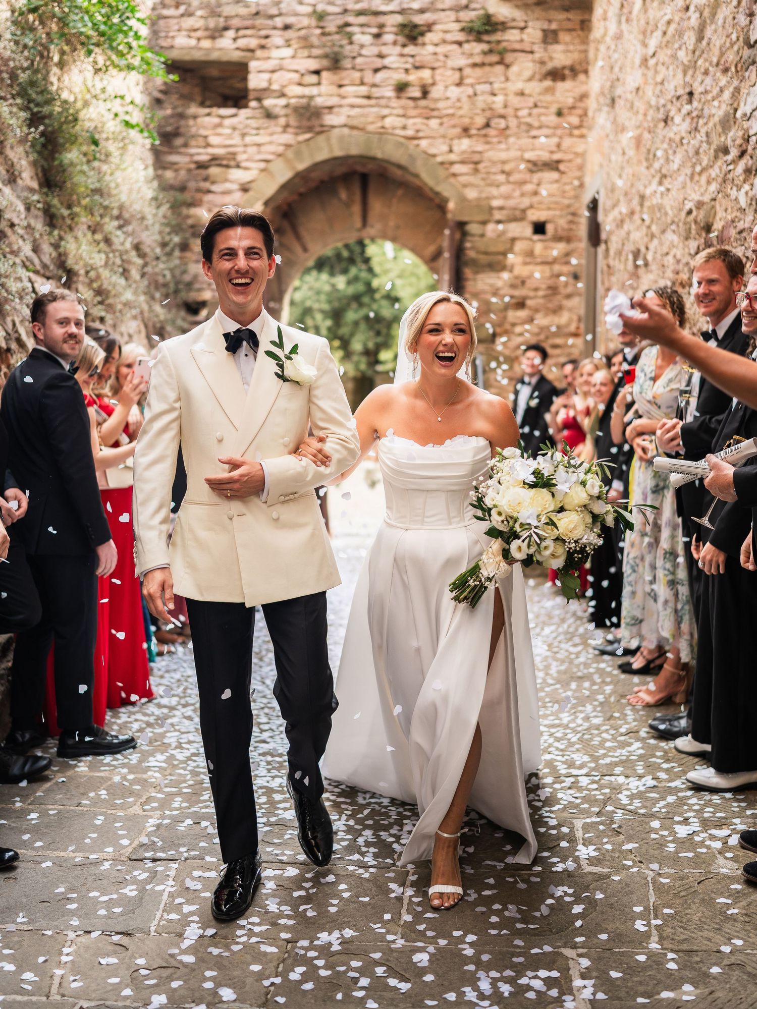 Joyful newlyweds walking through confetti shower in Italian castle courtyard after wedding ceremony.
