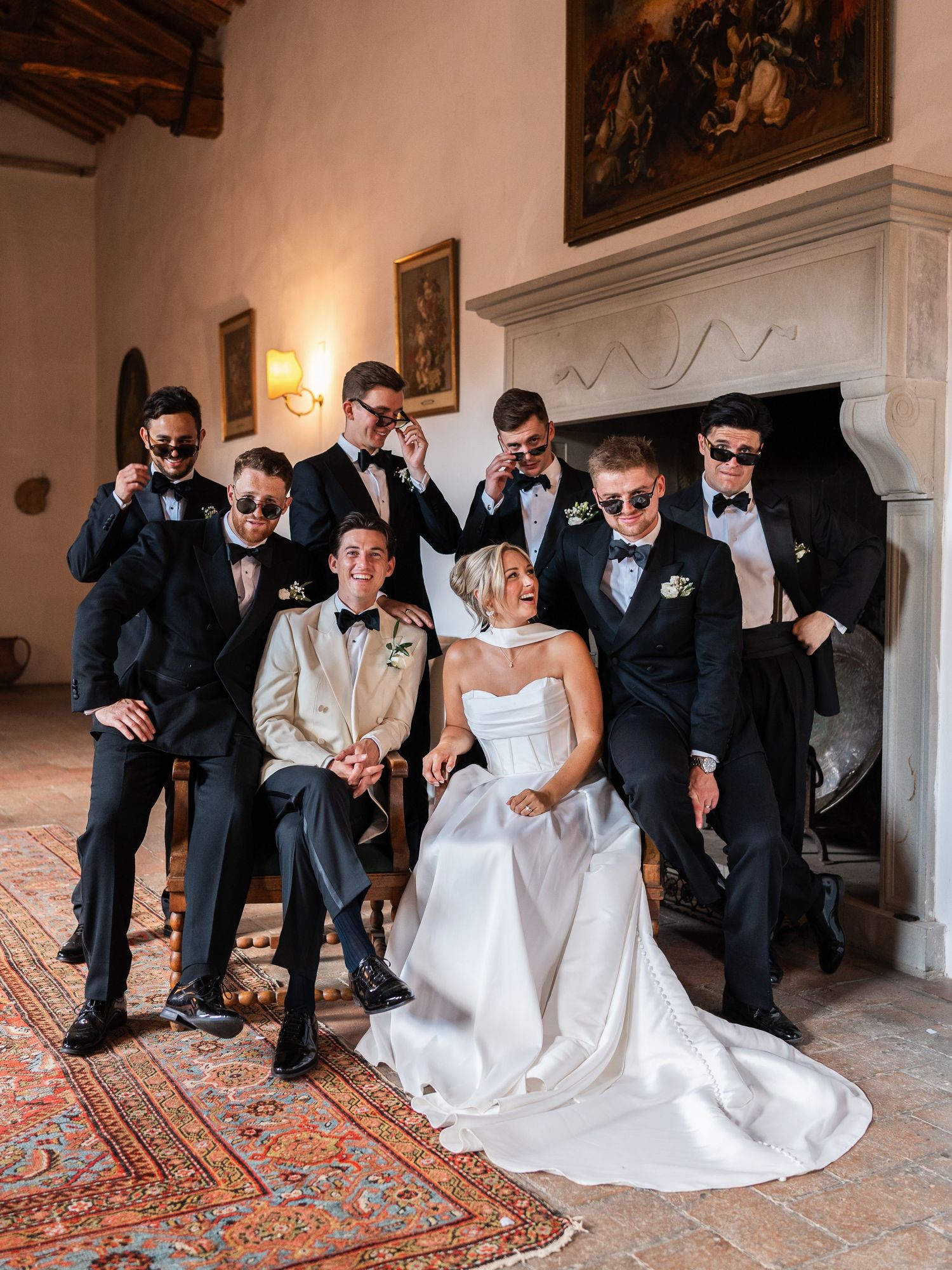Bride and groomsmen in black tuxedos posing with sunglasses by ornate fireplace in Italian villa.