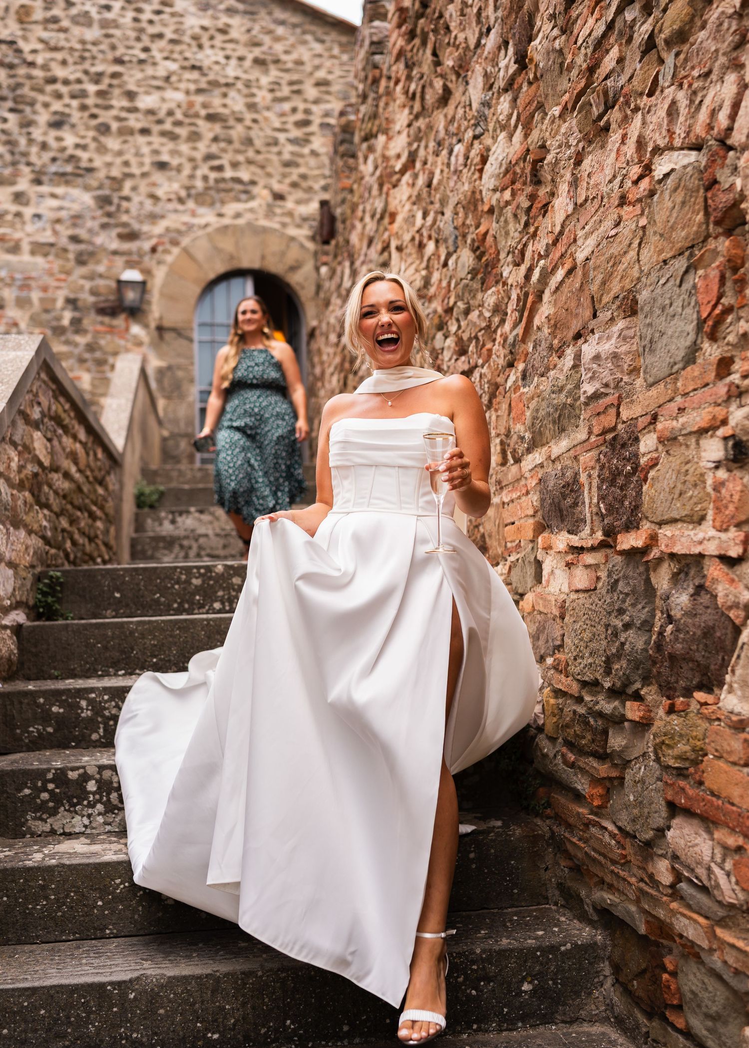 Joyful bride in white strapless gown holding champagne on stone castle steps.
