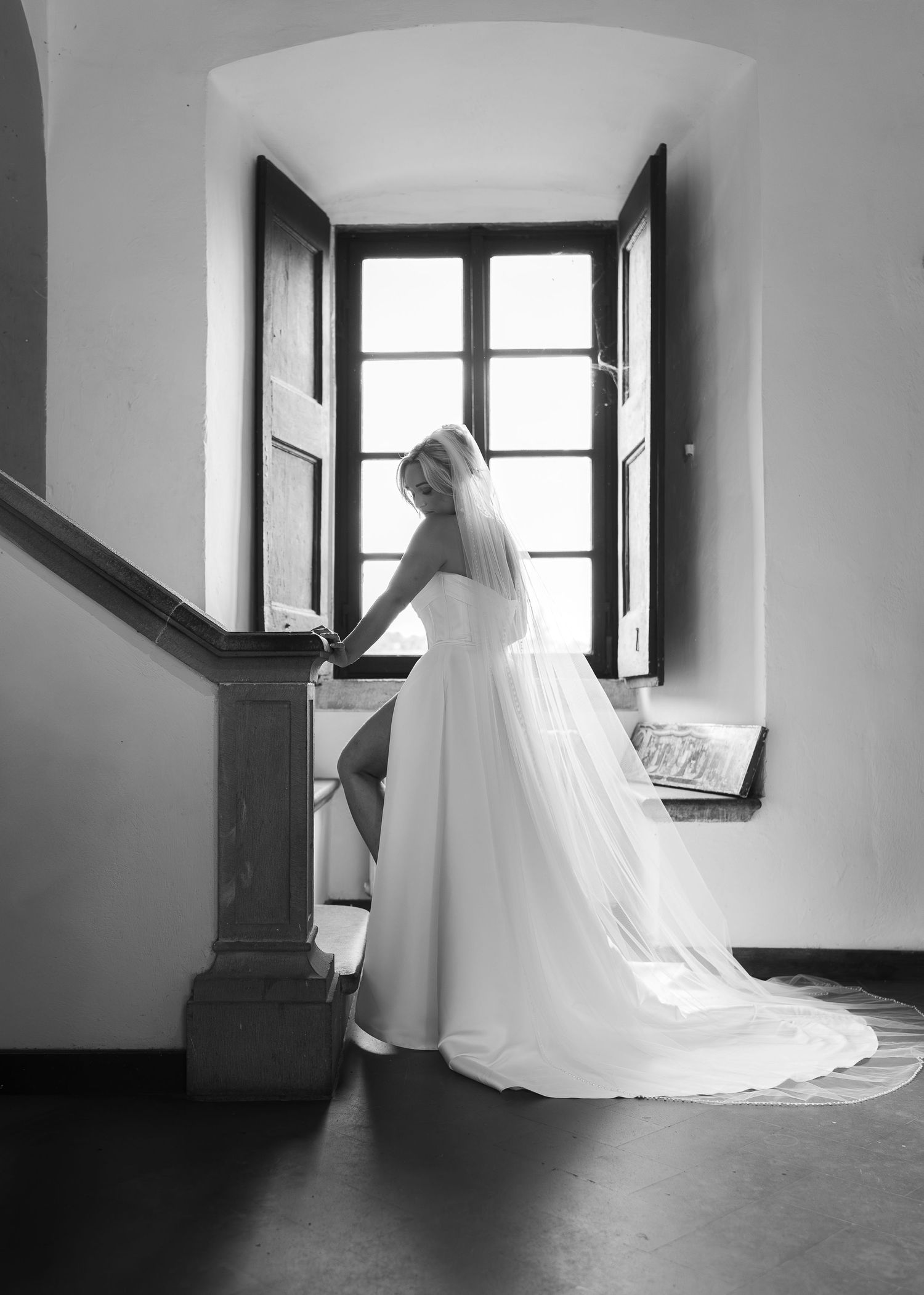 Elegant bride in a flowing white gown and veil standing by a window in a black and white photo.