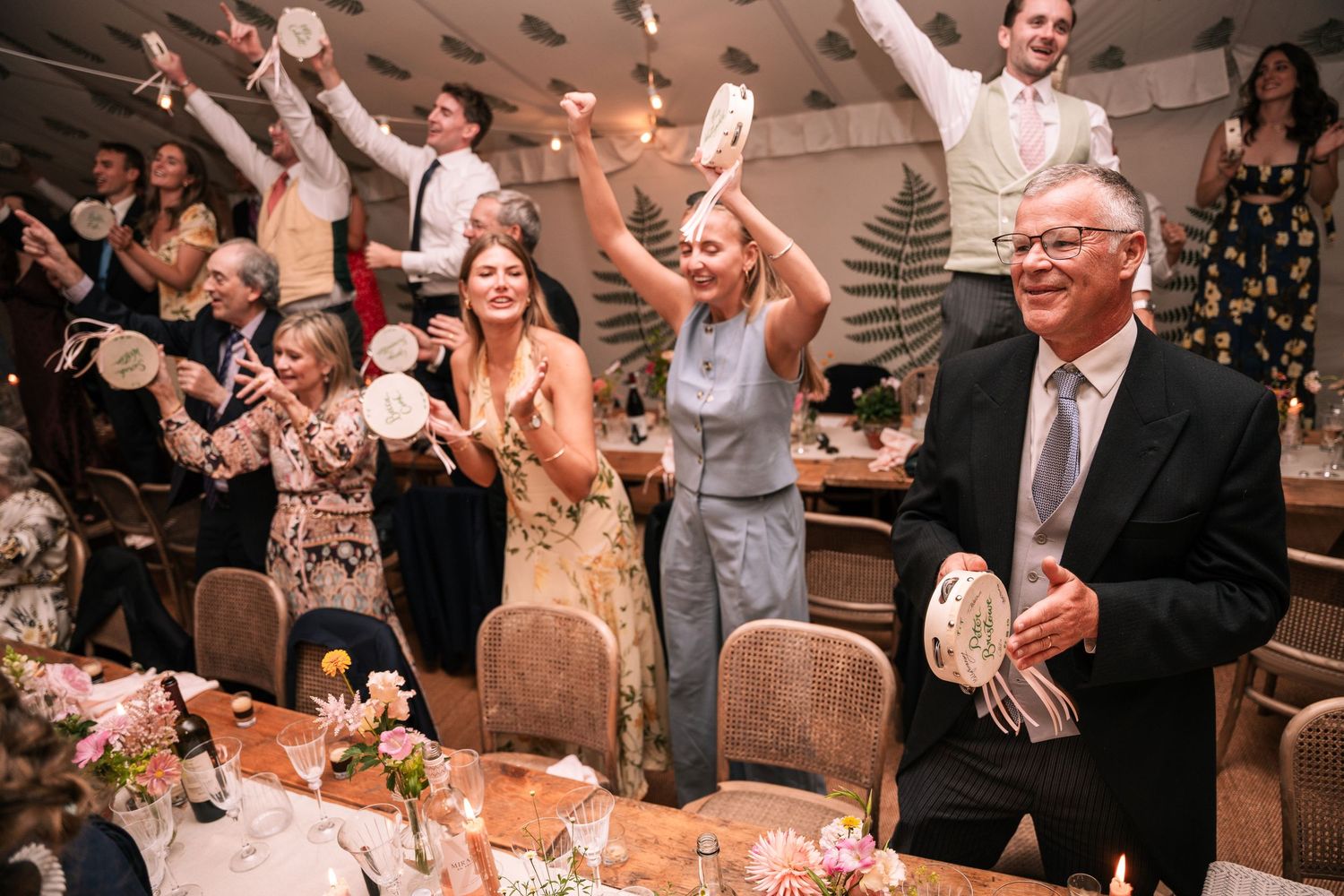 Cheerful wedding guests raising tambourines and celebrating at floral decorated reception tables.
