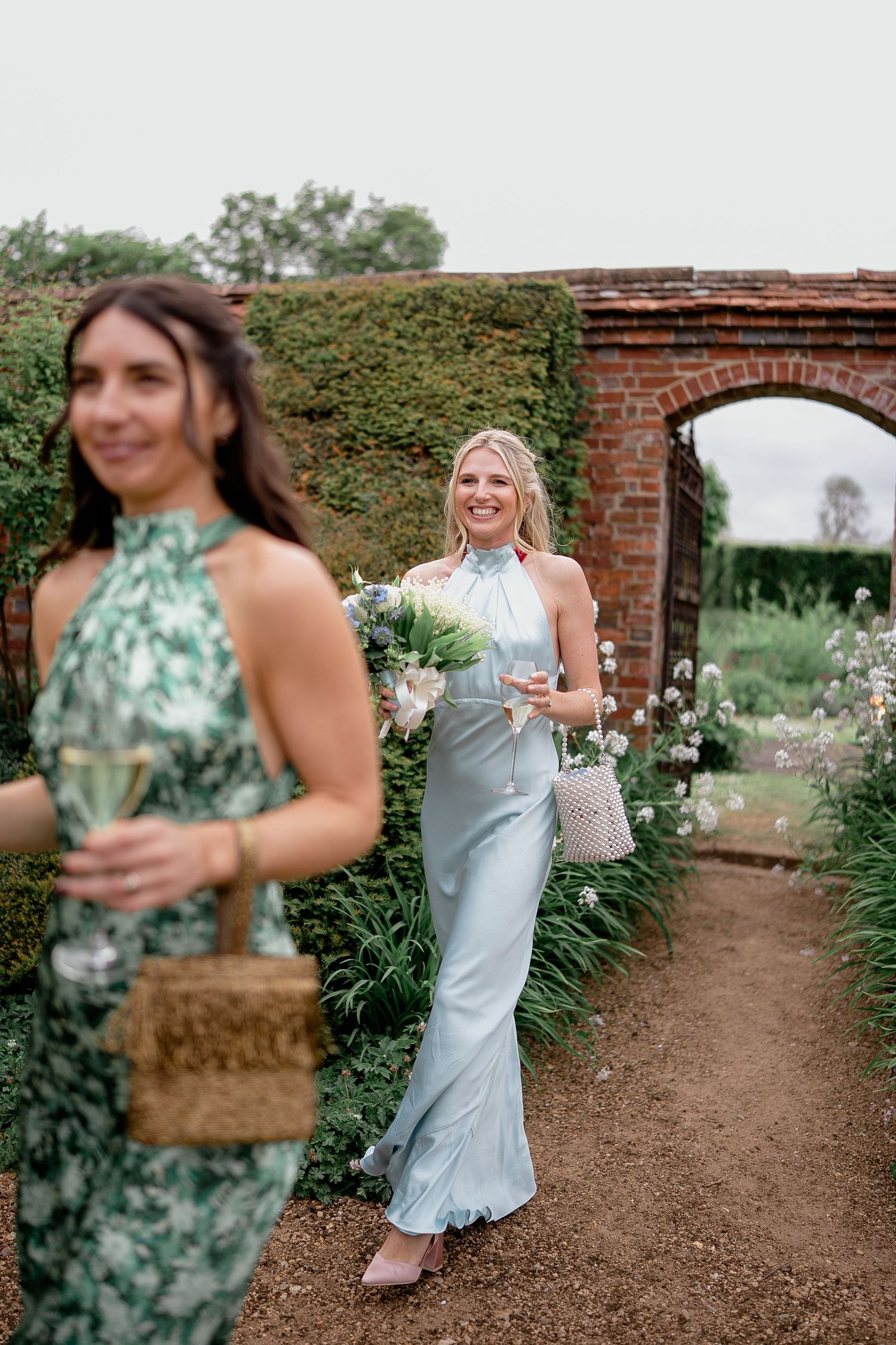 Bridesmaid in light blue satin halter dress holding bouquet walking through brick garden archway at wedding.