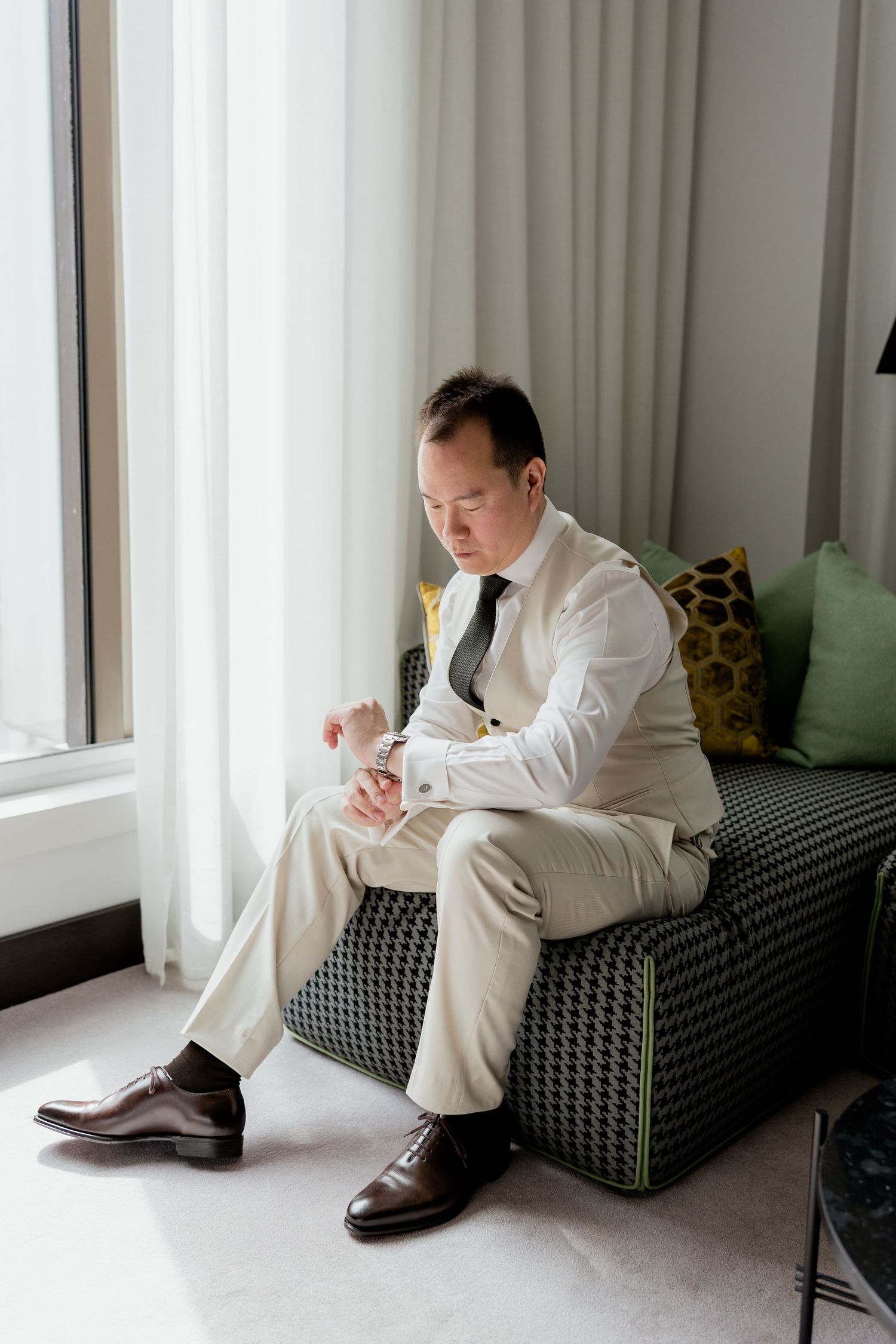 Groom in white shirt and beige suit adjusts cufflinks while seated by a sunlit window.