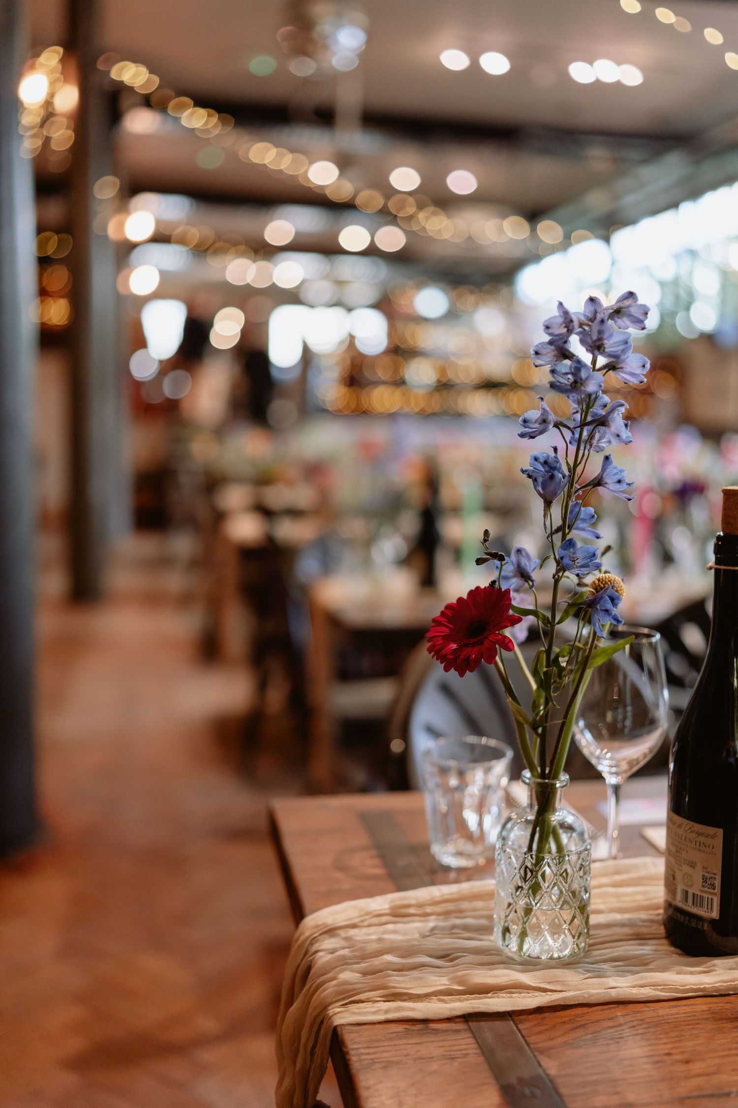 Crystal vase with blue delphiniums and red gerbera on a rustic wooden table in a softly lit venue.