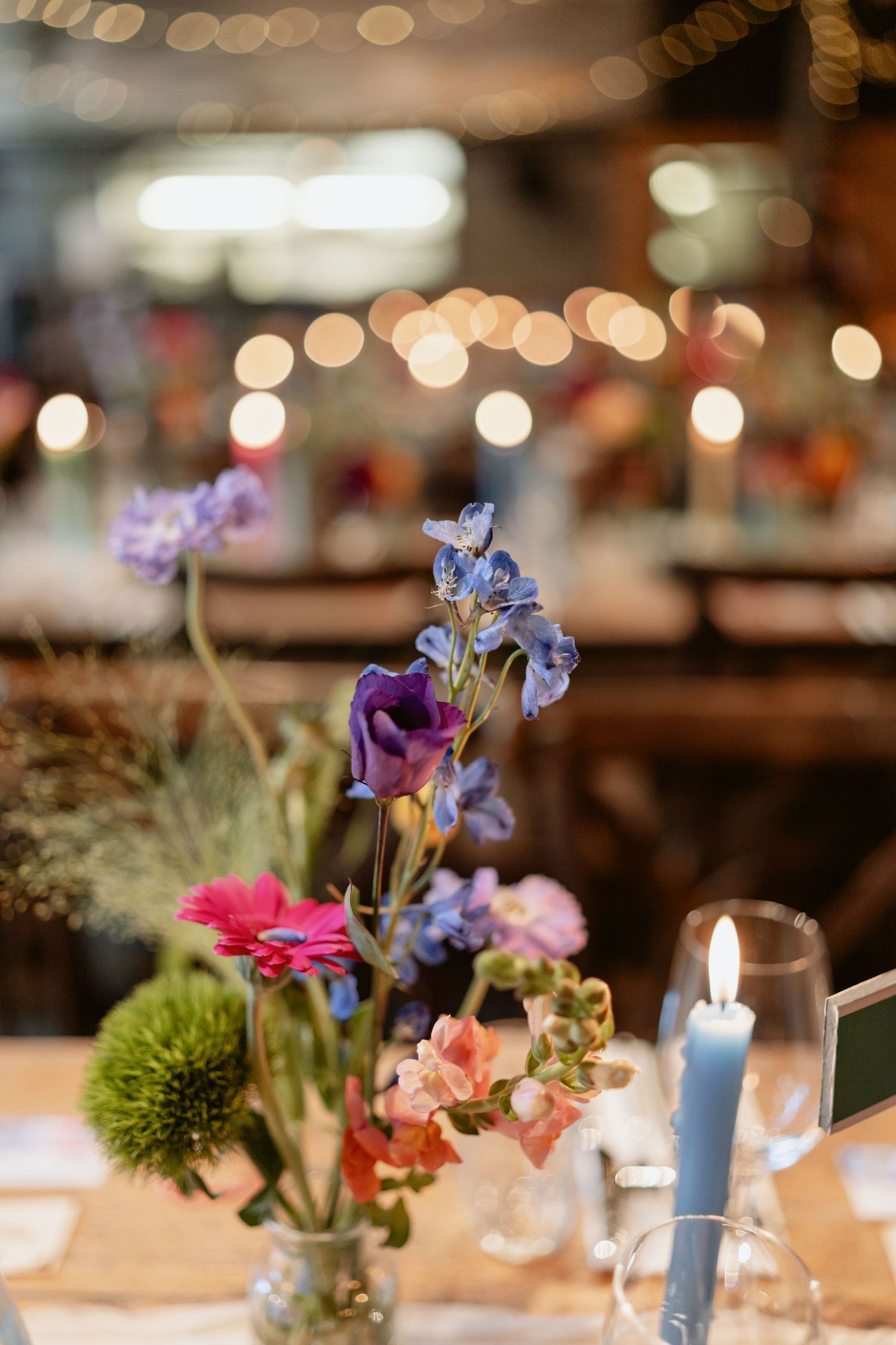 Colorful wildflower wedding centerpiece with blue candle and bokeh string lights in reception venue.