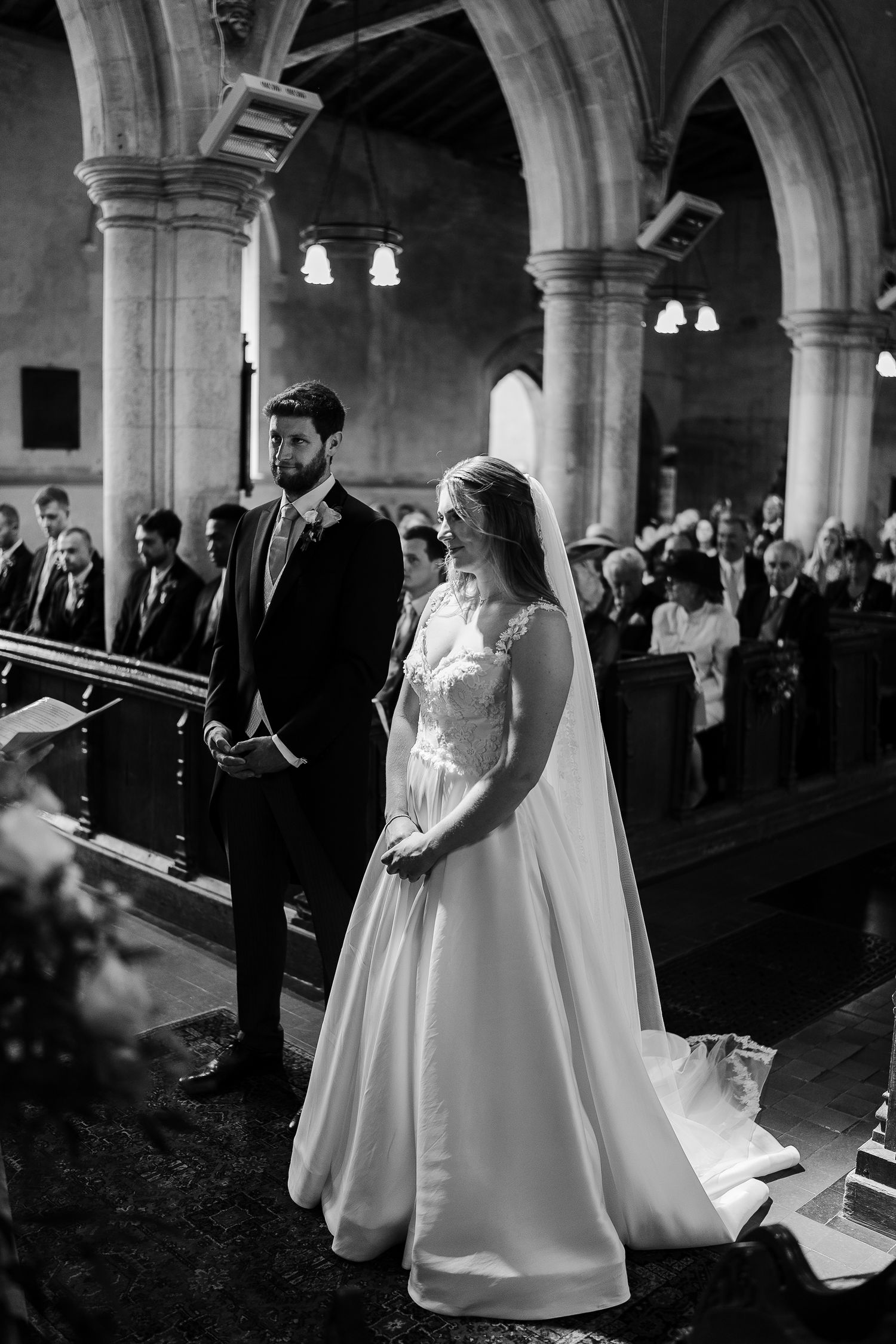 Black and white photo of bride in ballgown and groom standing at church altar during ceremony.