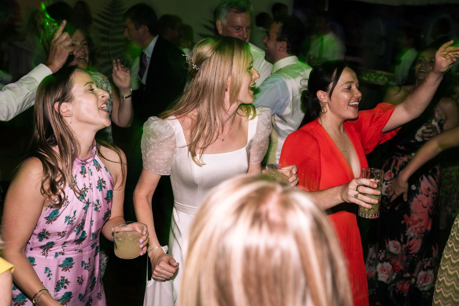 Three women in colorful dresses laughing and dancing at a wedding reception.