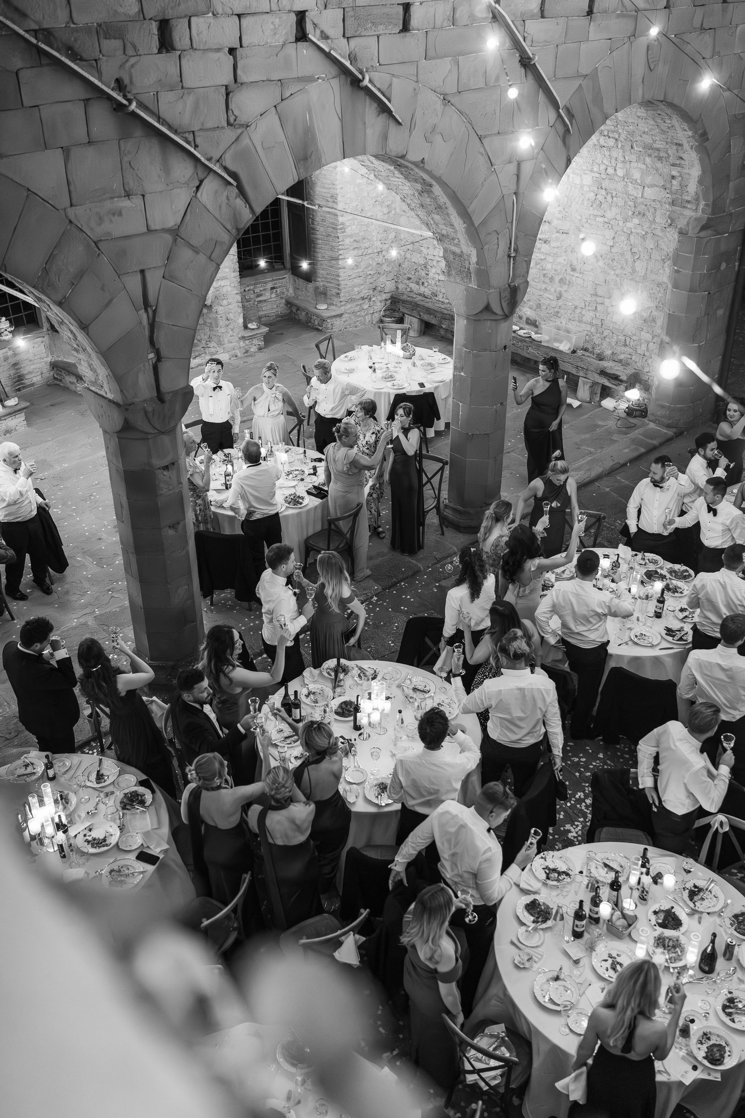 Black and white aerial view of wedding guests dining under arched stone ceilings with string lights.