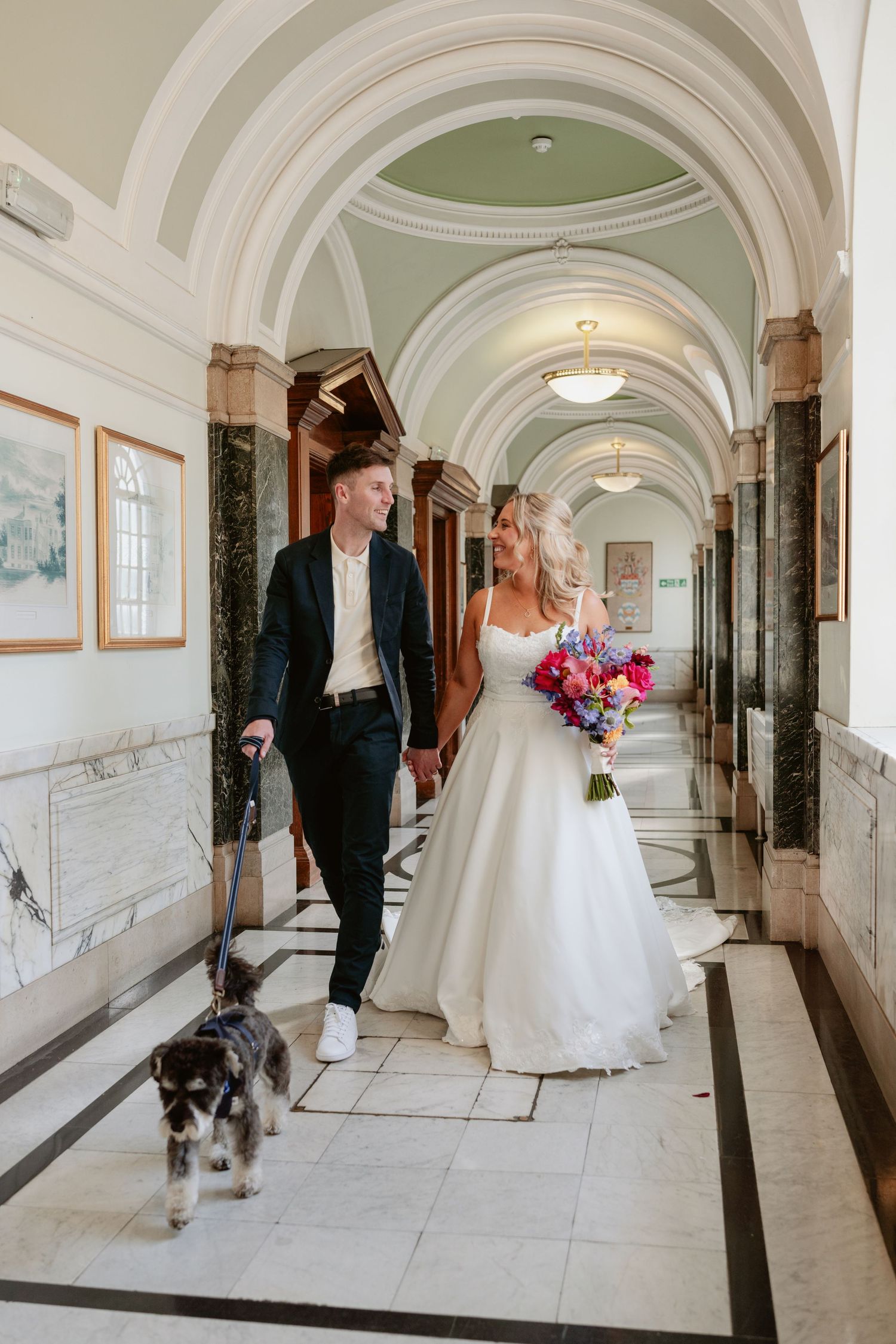 Bride and groom walking their dog through an elegant arched hallway holding a vibrant bridal bouquet.