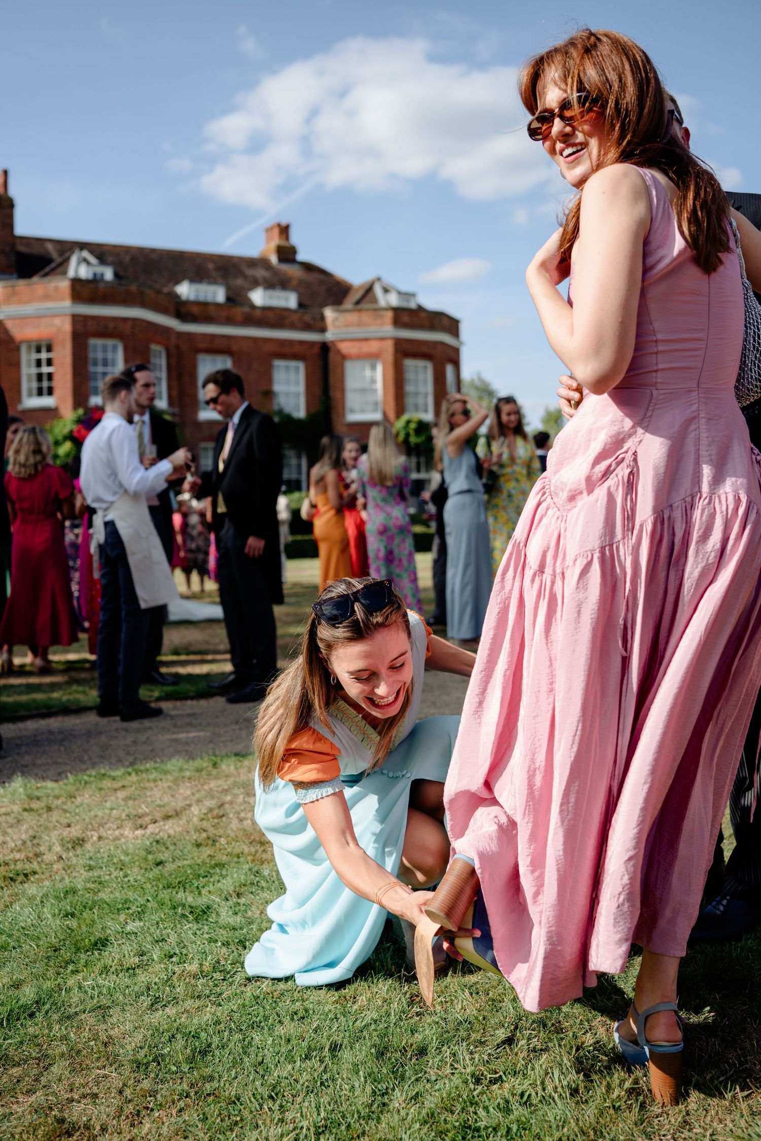 Laughing wedding guest in blue dress kneels on grass beside woman in pink at sunny reception.