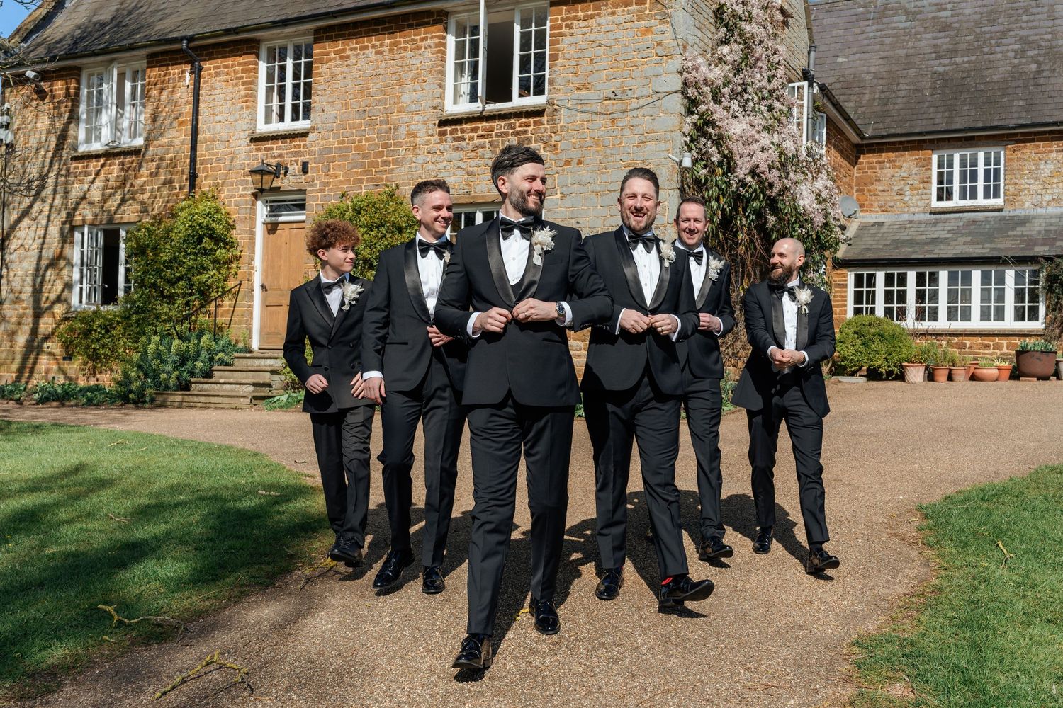 Groom and groomsmen in black tuxedos walk confidently outside a brick manor house.