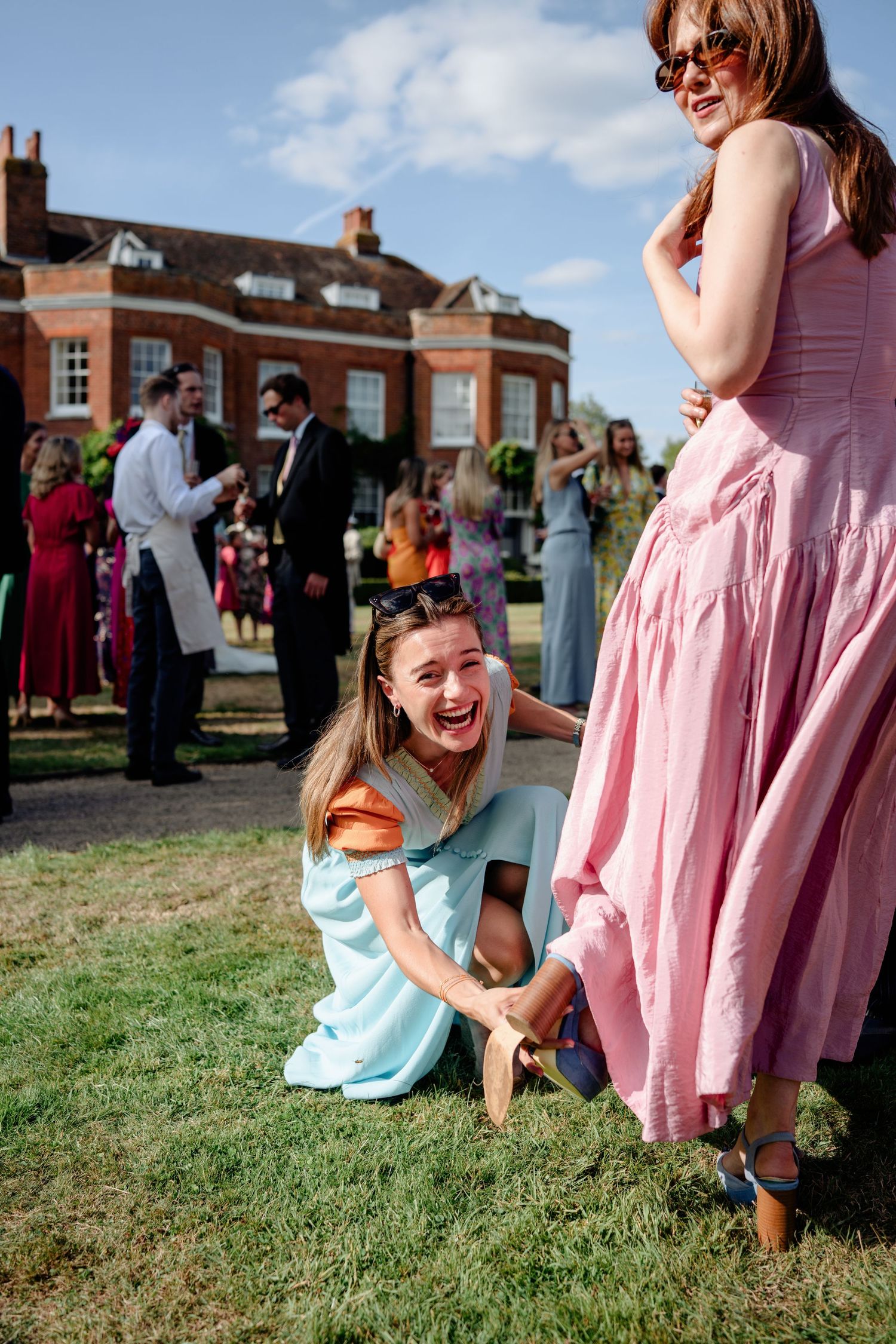 Joyful wedding guest in blue dress sits laughing on lawn beside woman in pink summer dress.