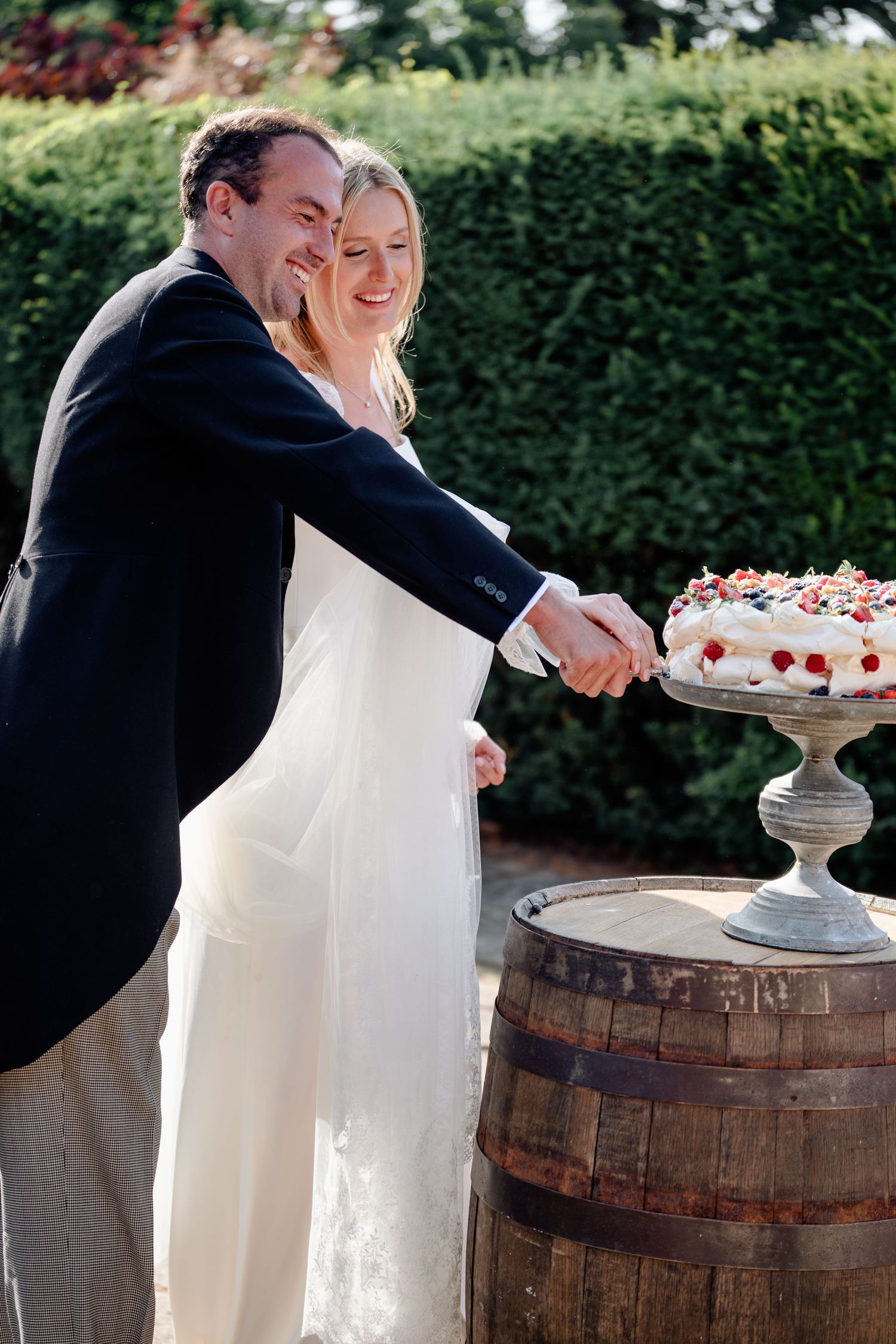 Newlyweds smiling as they cut their berry-topped wedding cake displayed on a rustic wooden barrel.