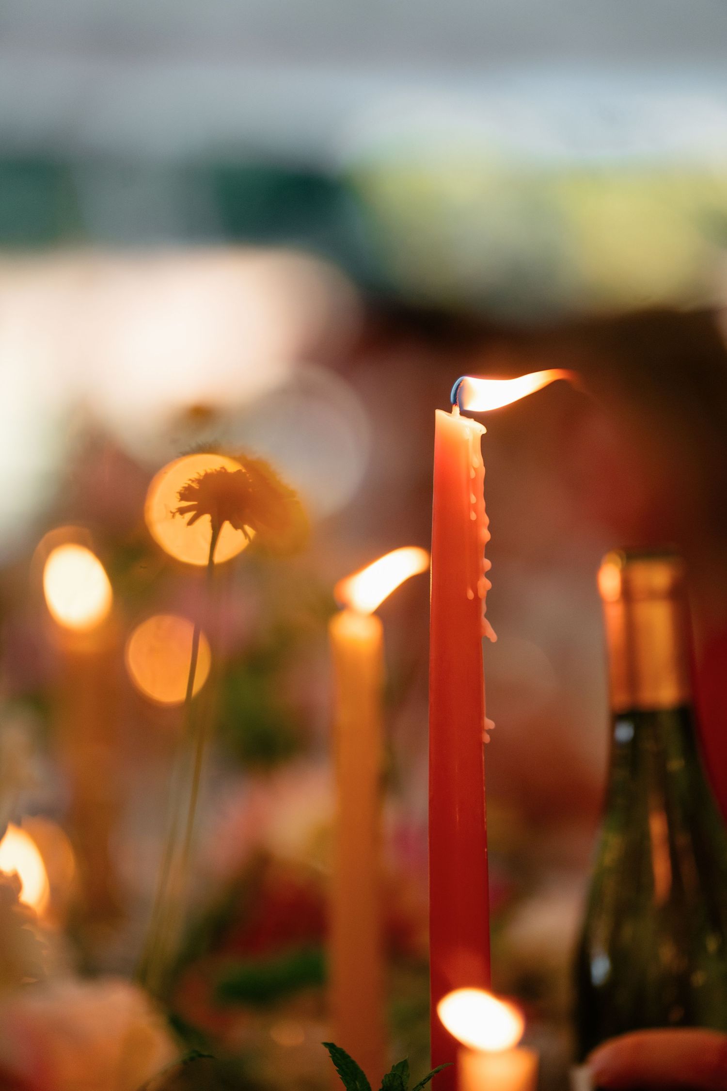 Lit red taper candle glowing warmly among flowers and bottles at an elegant wedding reception table.