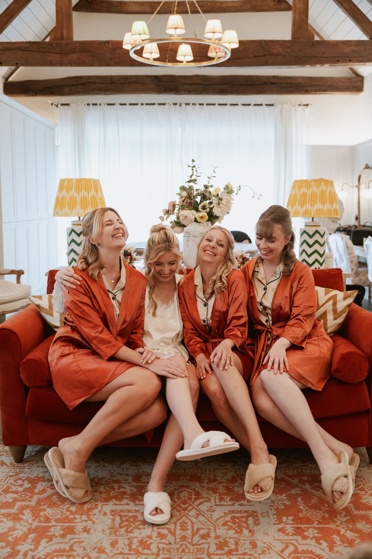 Four bridesmaids in rust orange satin robes sitting on a red sofa laughing together during wedding morning preparations.