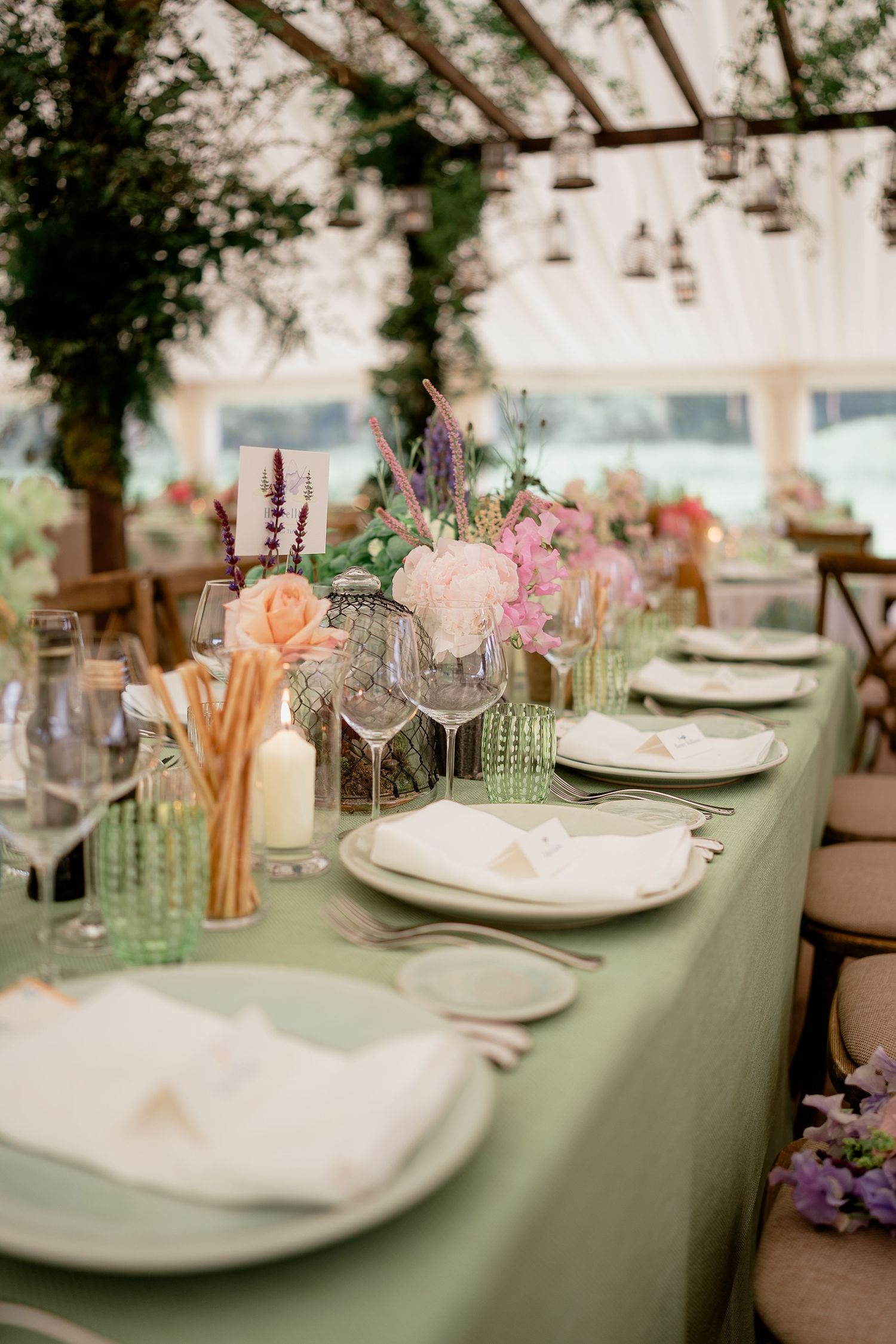 Elegant wedding table with sage green linen, pink floral centerpieces, candles, and crystal glassware in marquee.