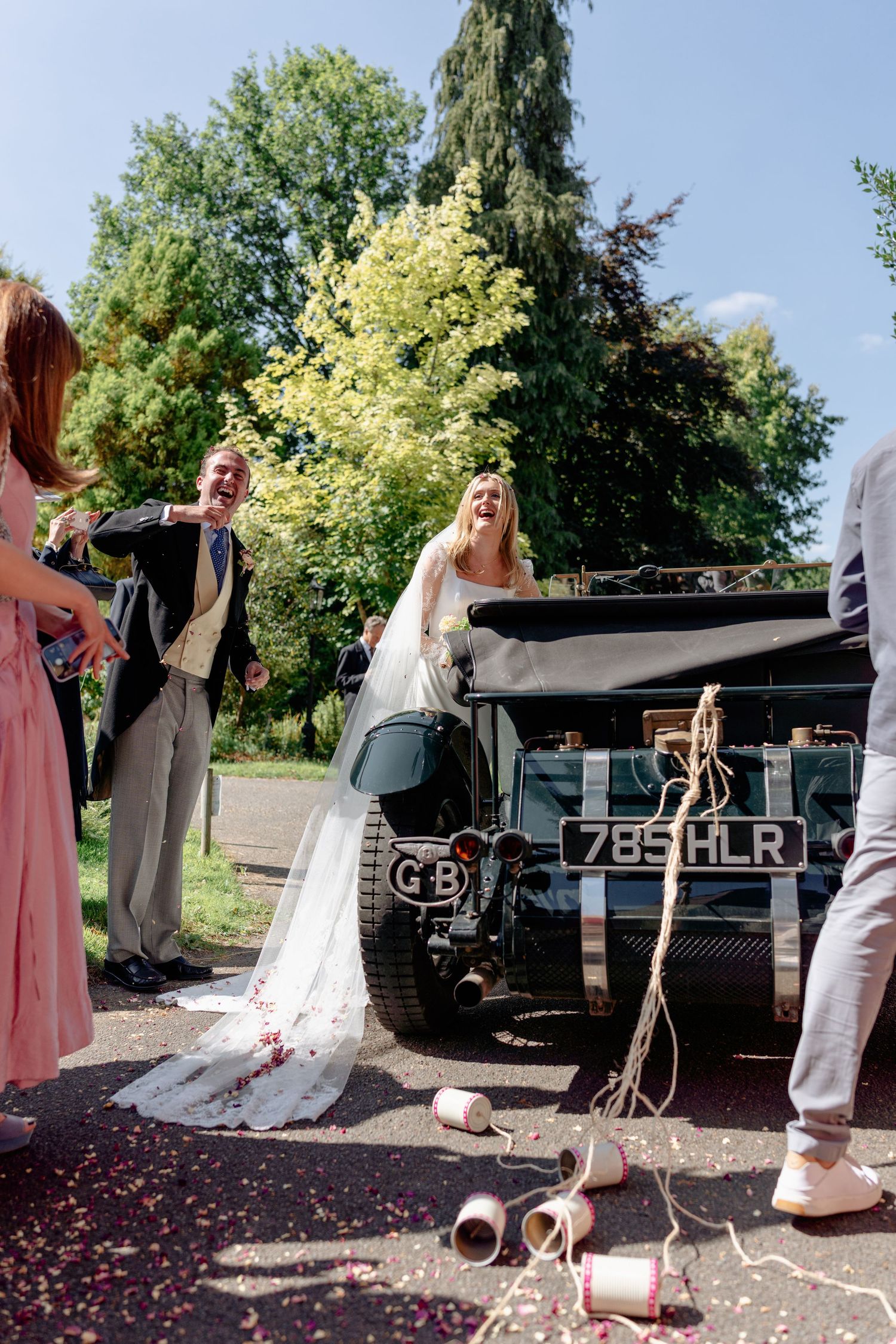 Laughing bride and groom pose by vintage GB Land Rover decorated with tin cans after wedding.