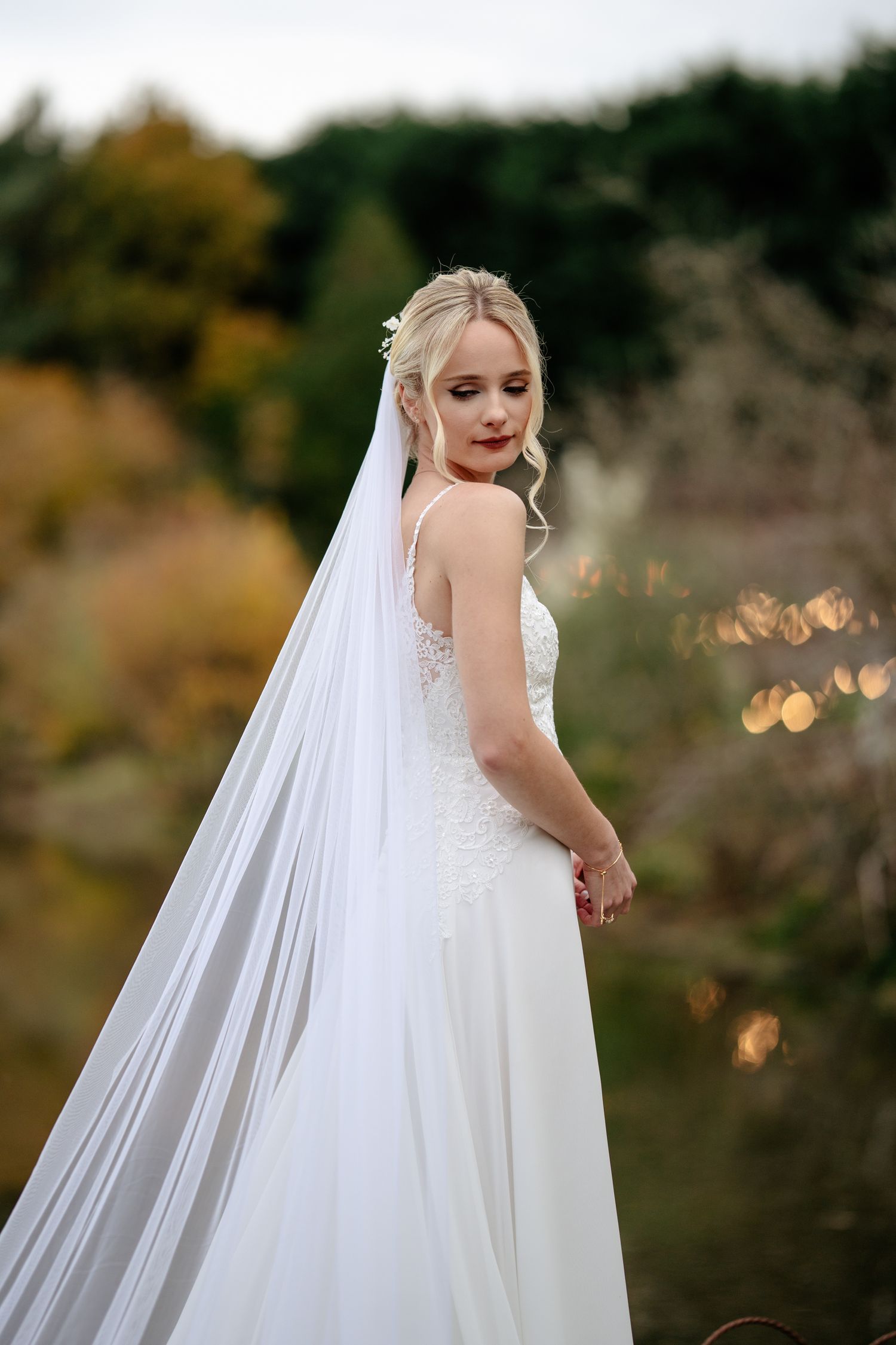 Elegant blonde bride in white lace gown and long veil poses in autumn garden.