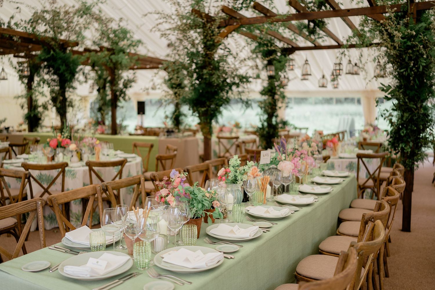 Elegant wedding reception marquee with sage green tablecloths, wooden cross-back chairs, and lush floral centerpieces.