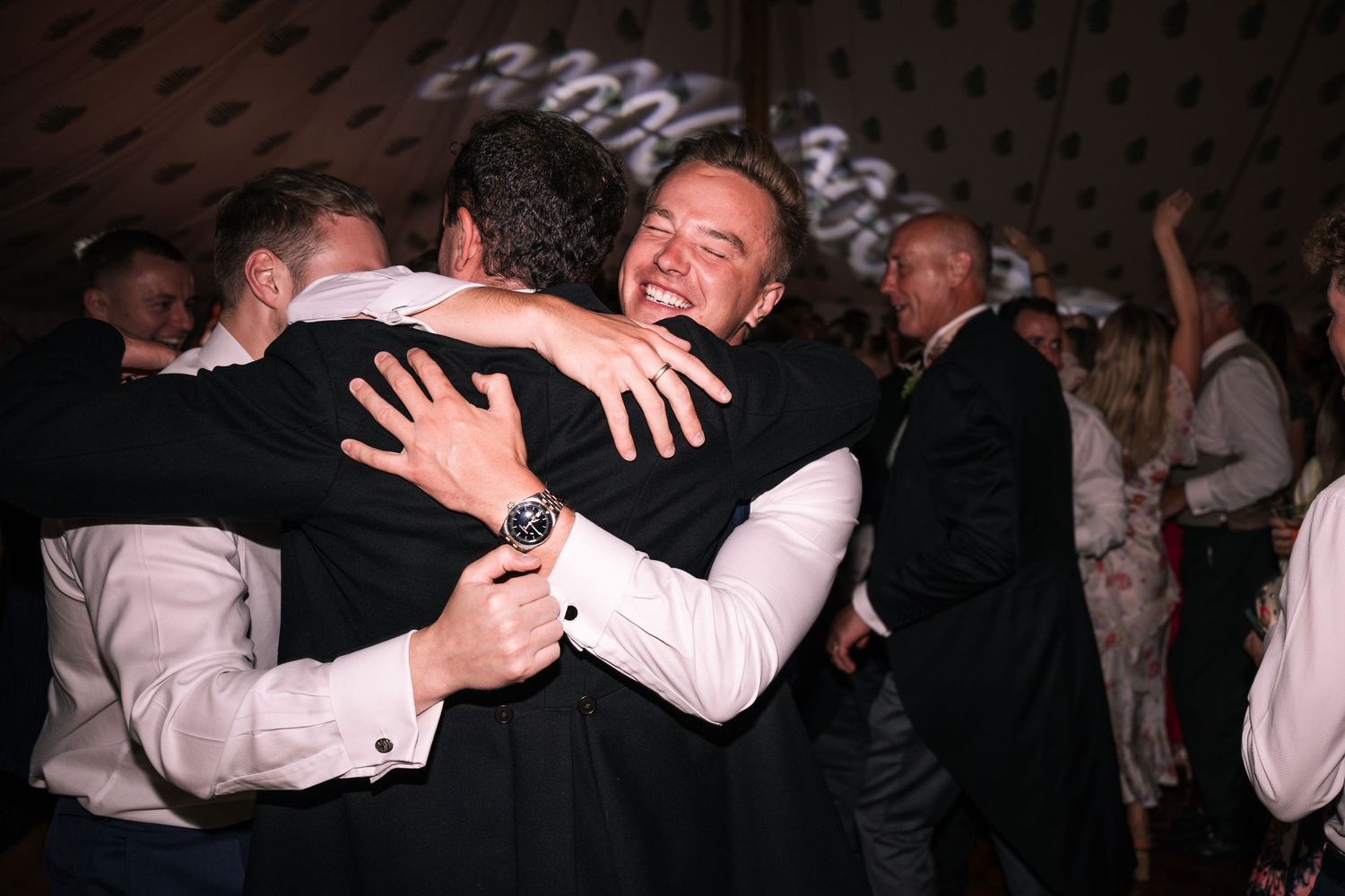 Two men sharing warm embrace on crowded wedding reception dance floor.