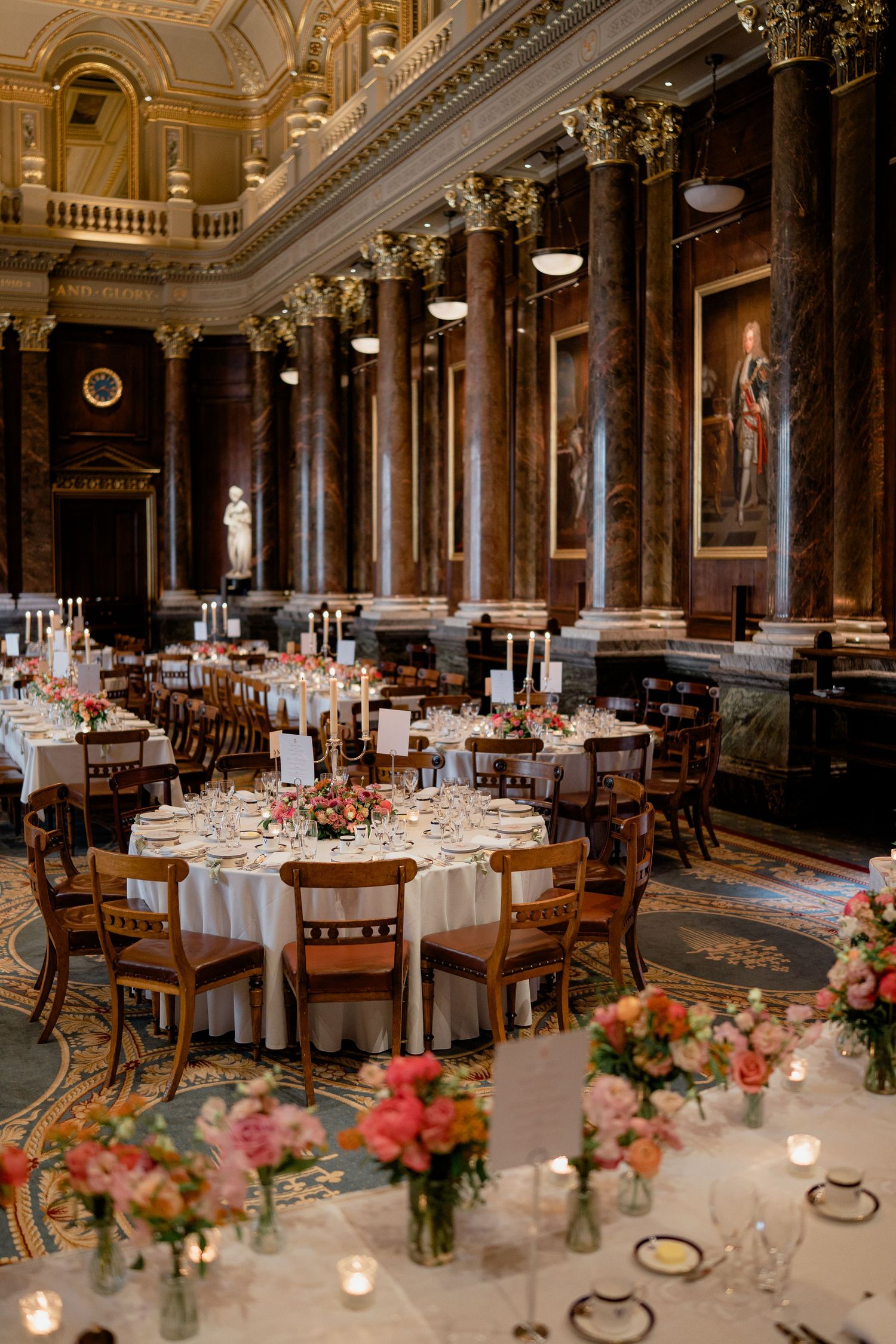 Grand wedding reception hall with marble columns, round tables, and pink floral centerpieces.