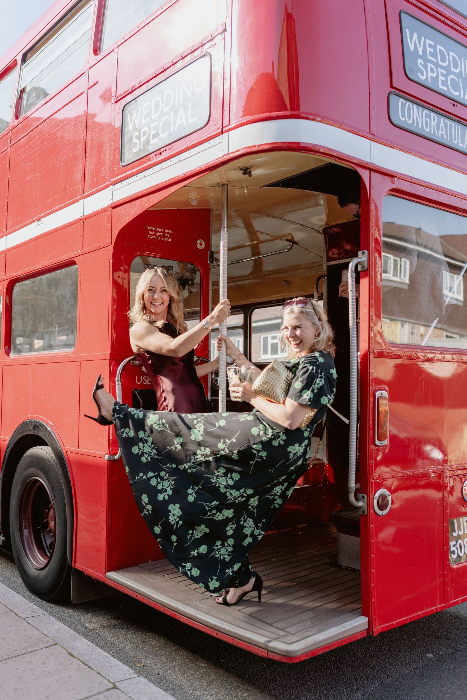Two elegantly dressed women posing and smiling on red London double-decker wedding bus.
