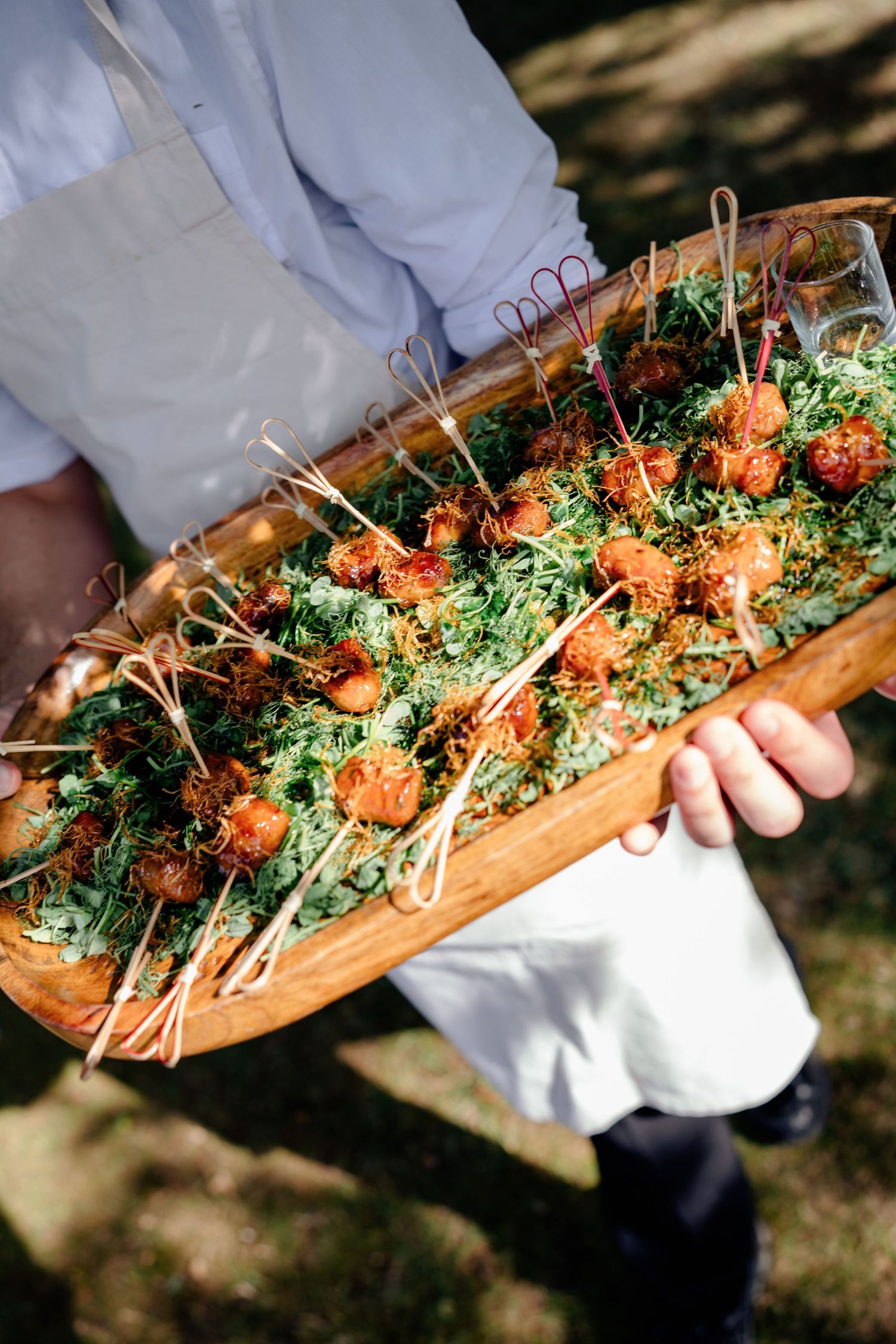 Caterer holds large wooden tray of golden fried canapés with decorative skewers at outdoor event.