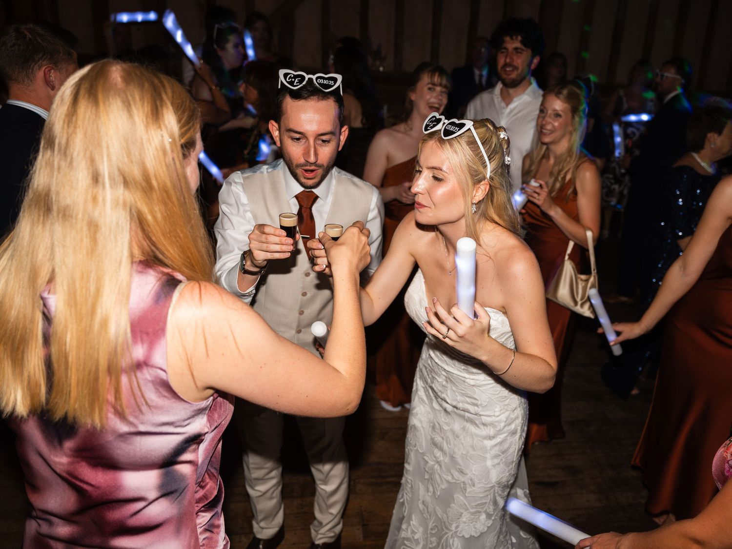Bride and groom celebrate with guests holding glow sticks on the wedding reception dance floor.