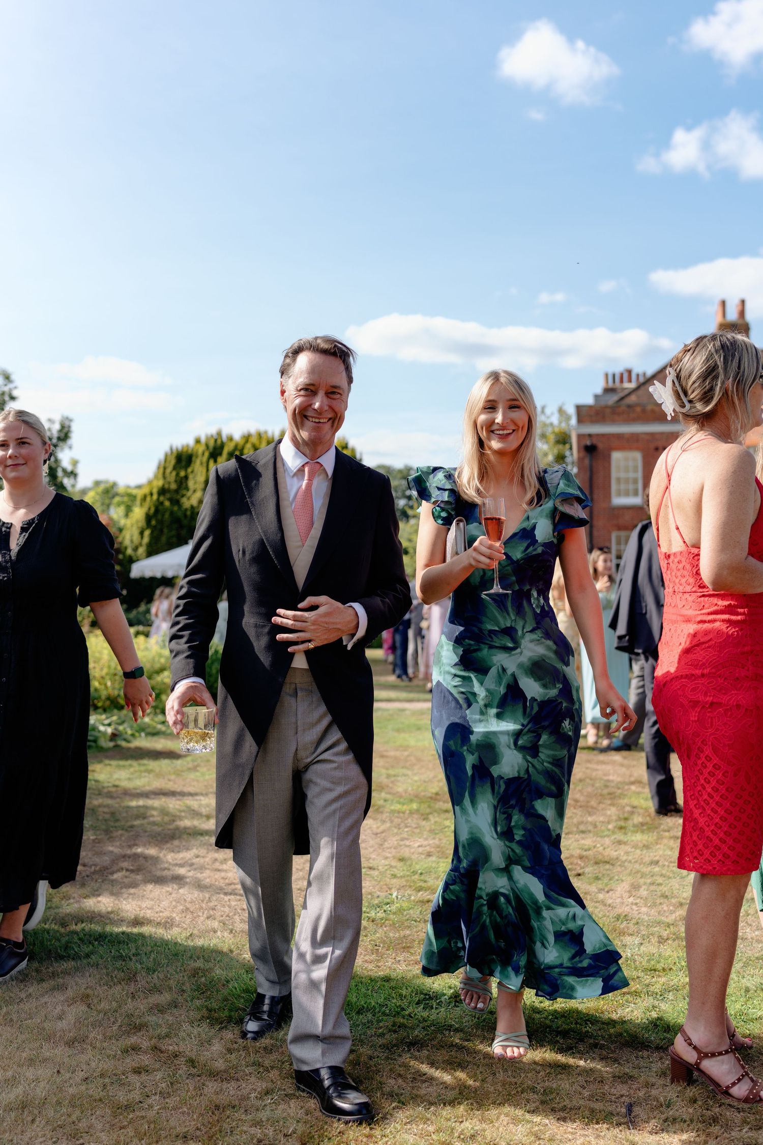 Elegantly dressed wedding guests walk across sunny garden grounds of a grand English manor house.