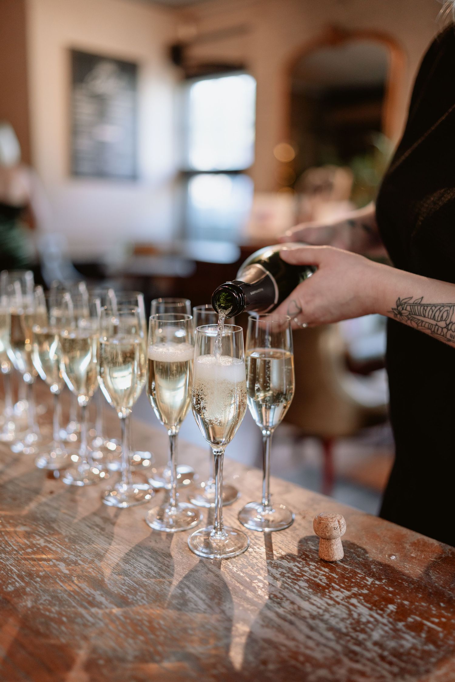 Tattooed bartender pouring champagne into a row of flute glasses on a rustic wooden bar.