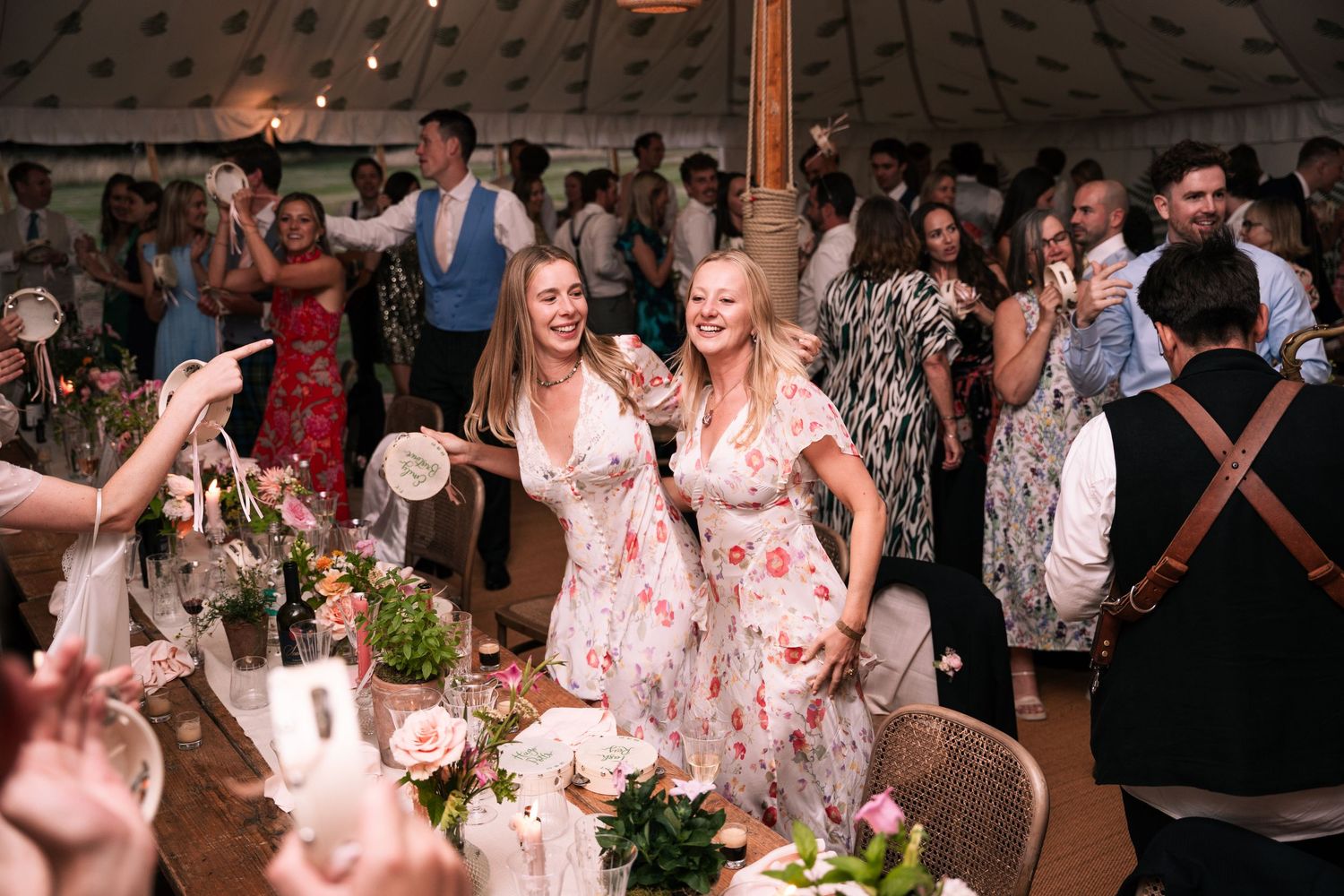 Two bridesmaids in floral dresses dancing joyfully at elegant tented wedding reception.