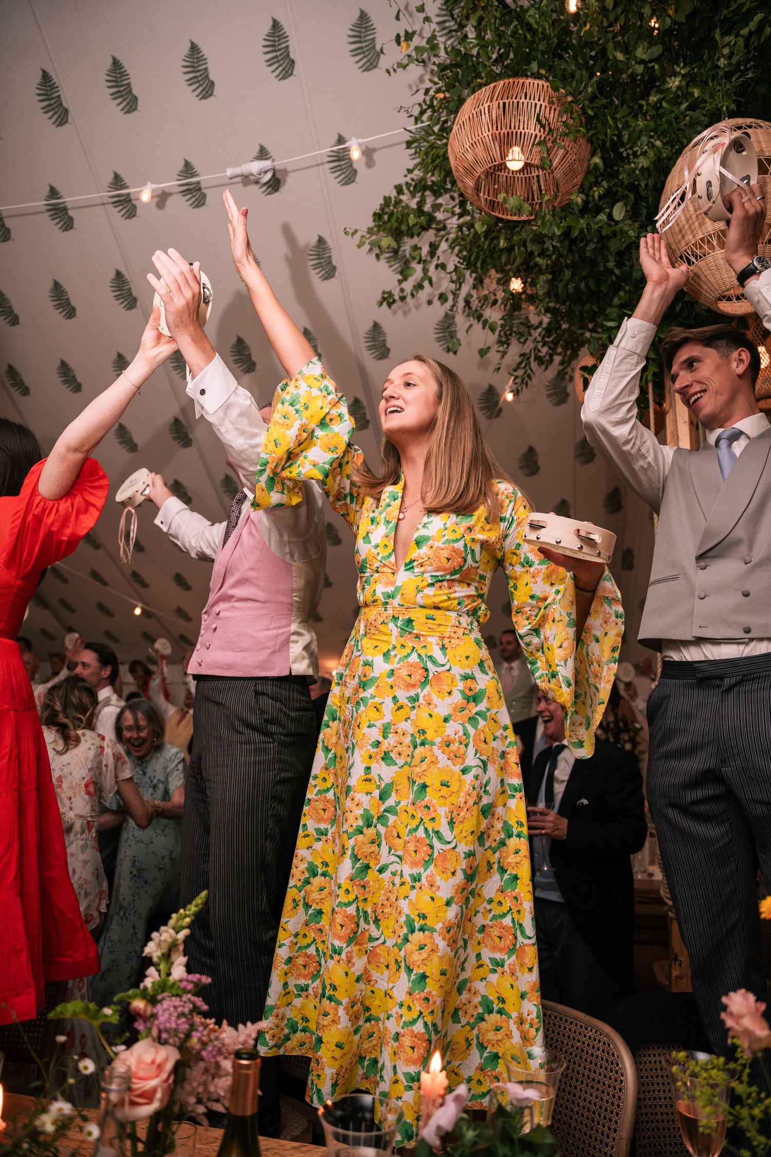 Elegant woman in yellow floral dress dancing with raised arms at wedding reception.