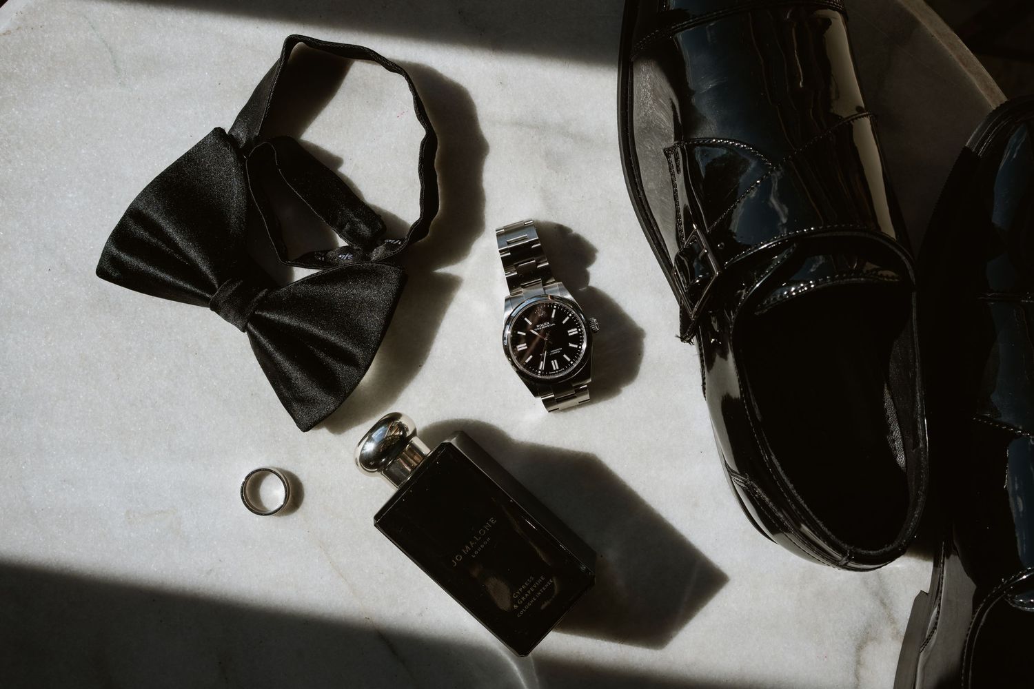 Flat lay of groom wedding accessories including black bow tie, watch, ring, cologne, and patent shoes.