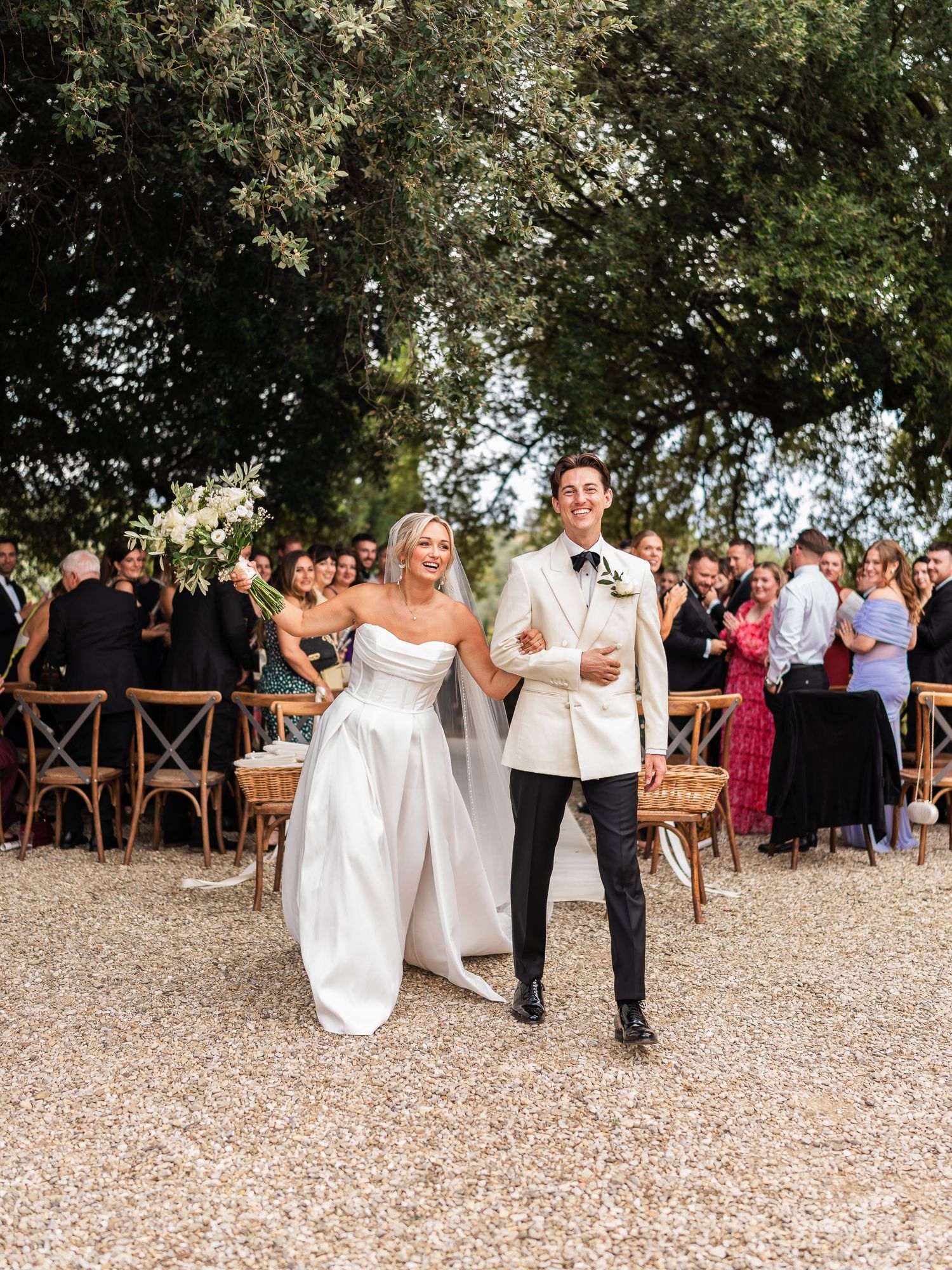 Bride in strapless gown and groom in cream tuxedo walk arm in arm down outdoor ceremony aisle.